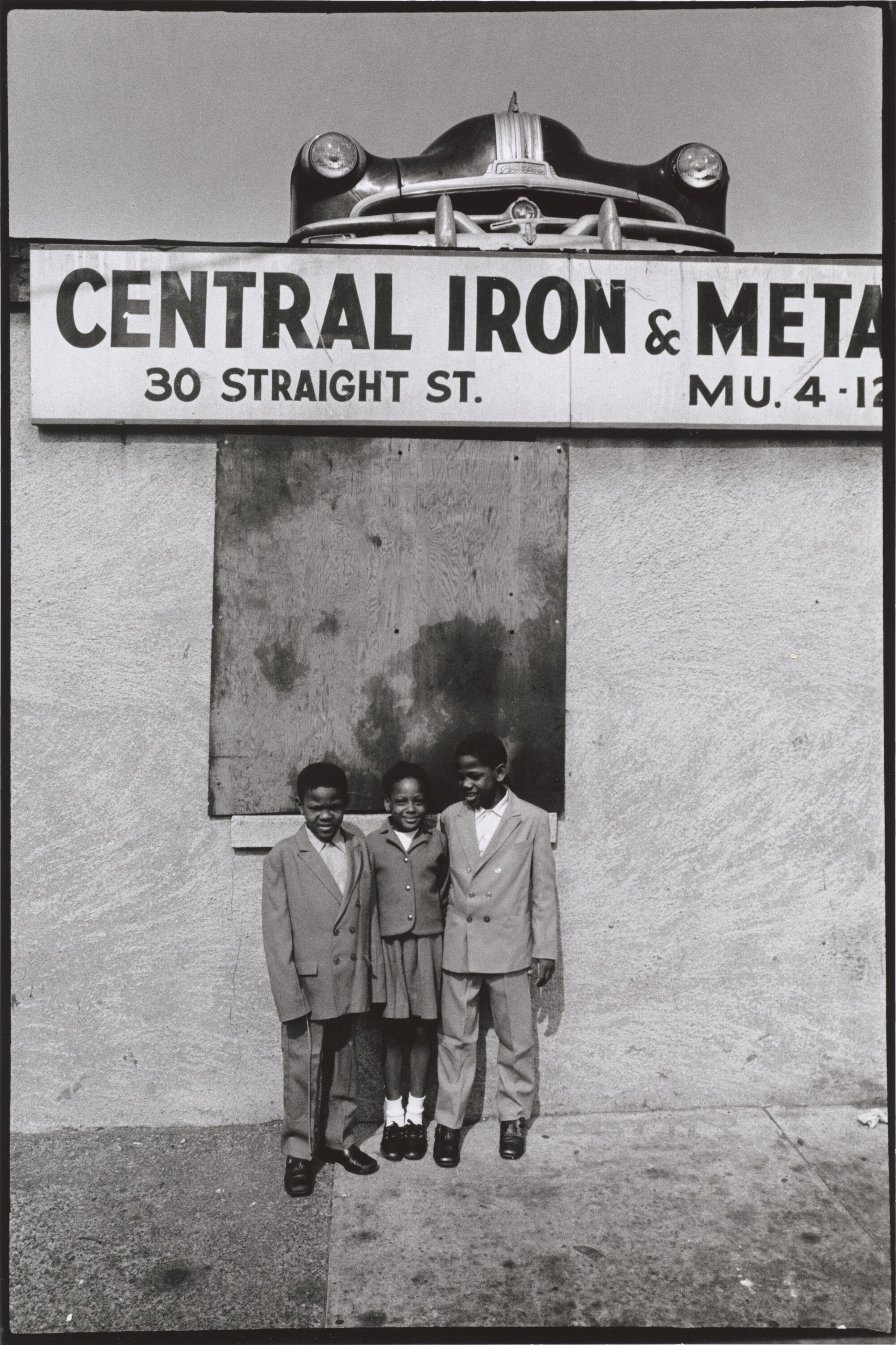 Three children standing together in front of a sign for a metal shop.