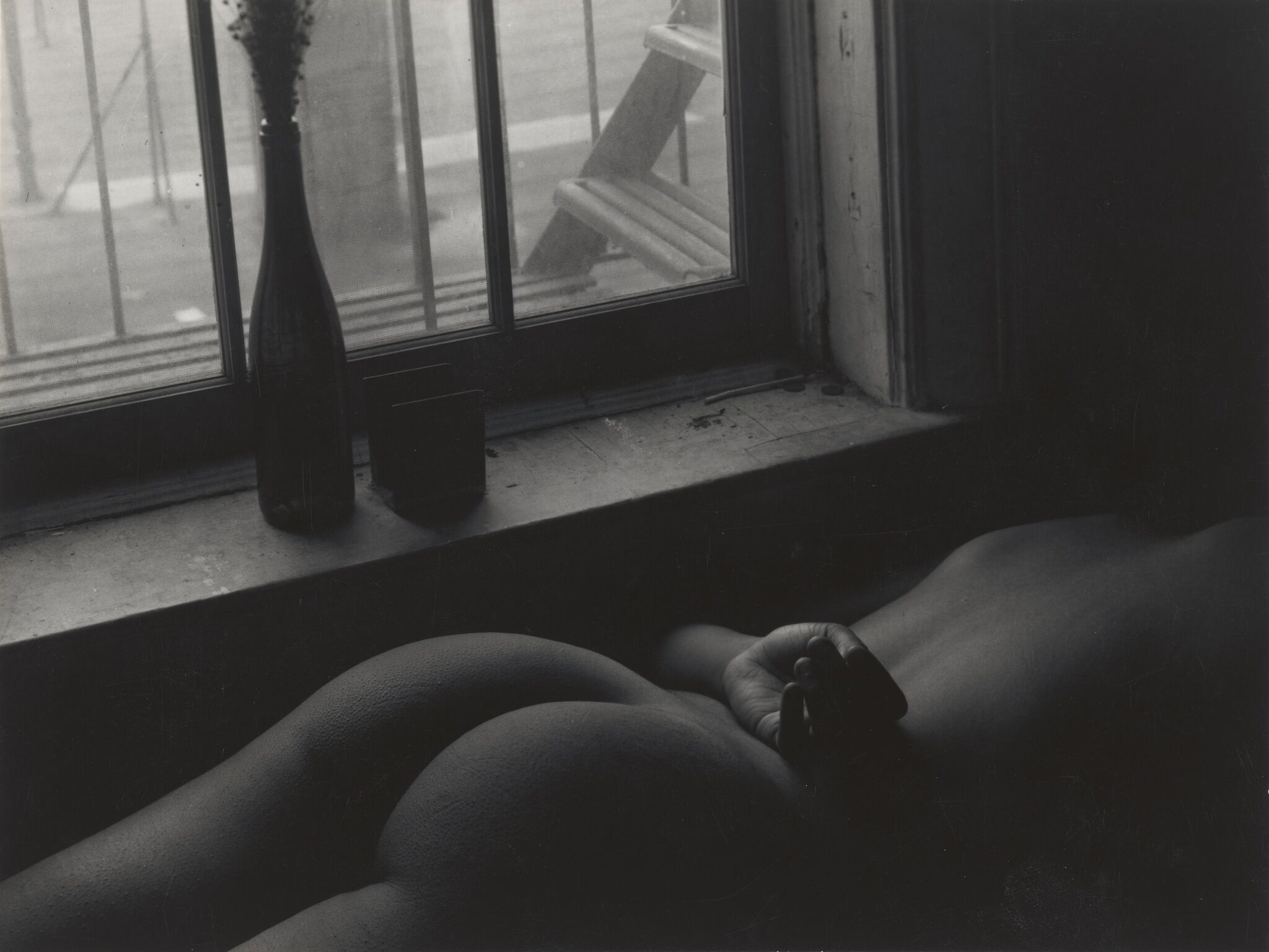 The backside of a nude person lying face down with their hand on their back, next to a window.