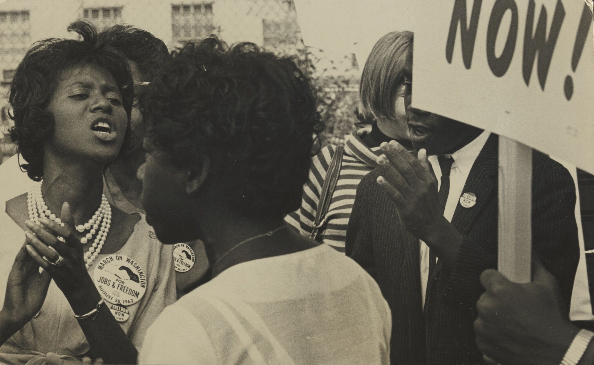 A woman within a group of people speaking out loud alongside a partial view of a person holding a picket sign.