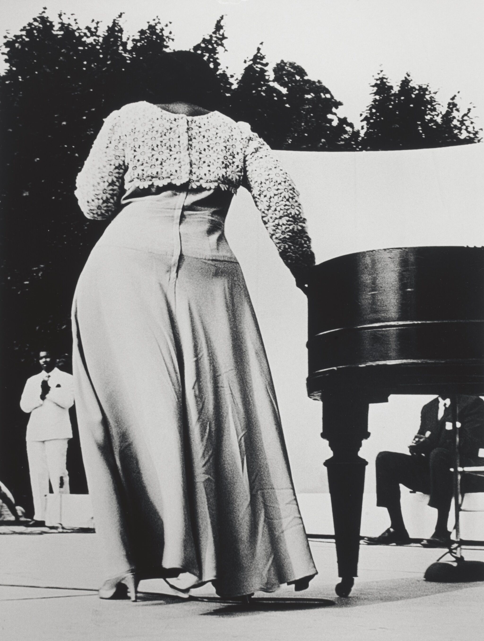 A woman with her back turned standing outdoors in a dress and high-heeled shoes.