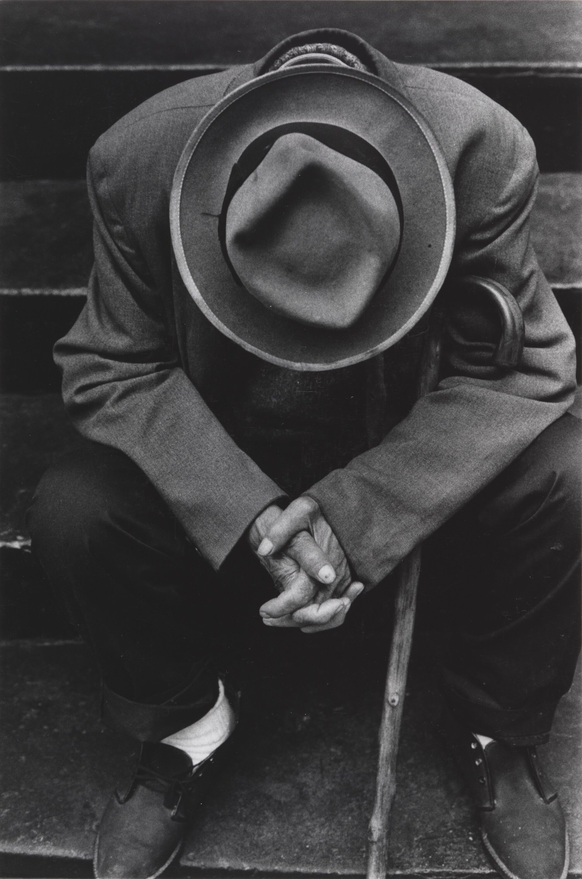 A person wearing a hat sitting with their head down and their hands clasped together.