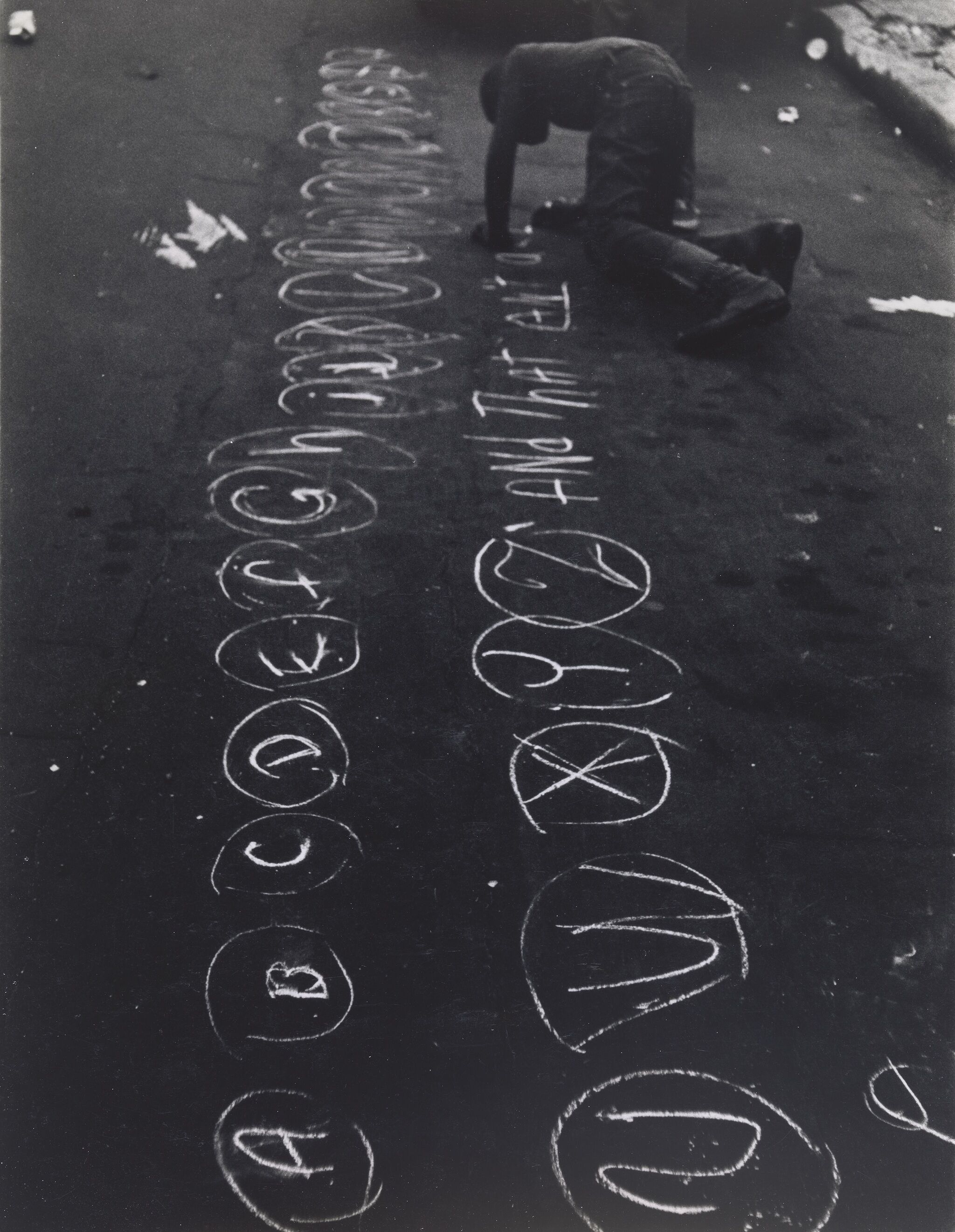 Letters of the alphabet handwritten on pavement with a person crouched and writing on the ground.
