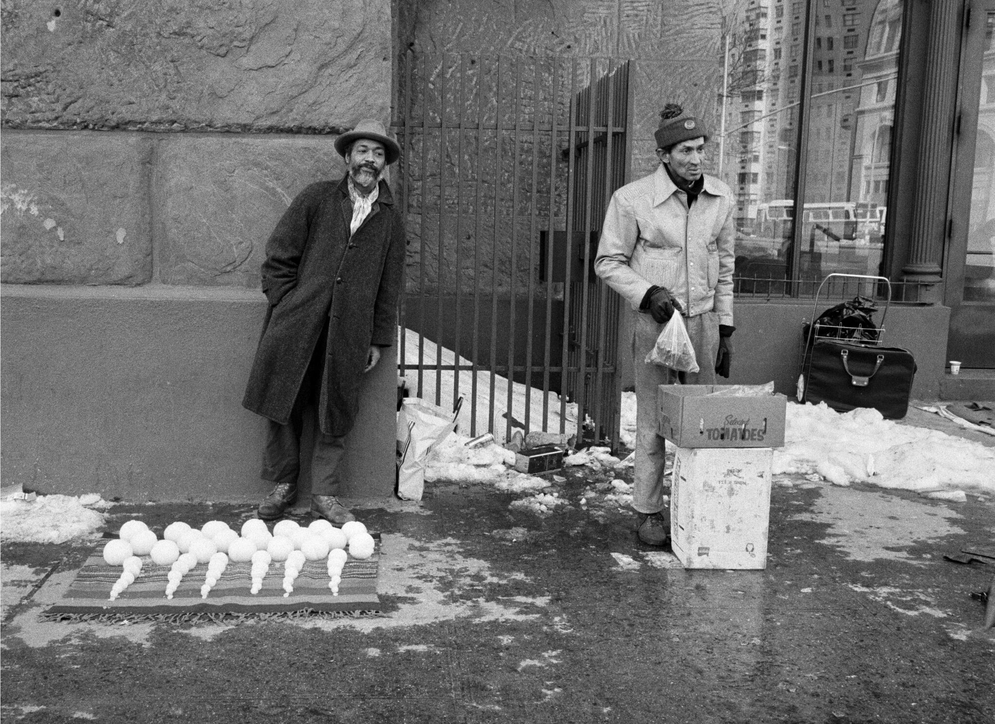 Two men standing on the street. The man on the left leans against the wall while the other stands behind a short pile of boxes.