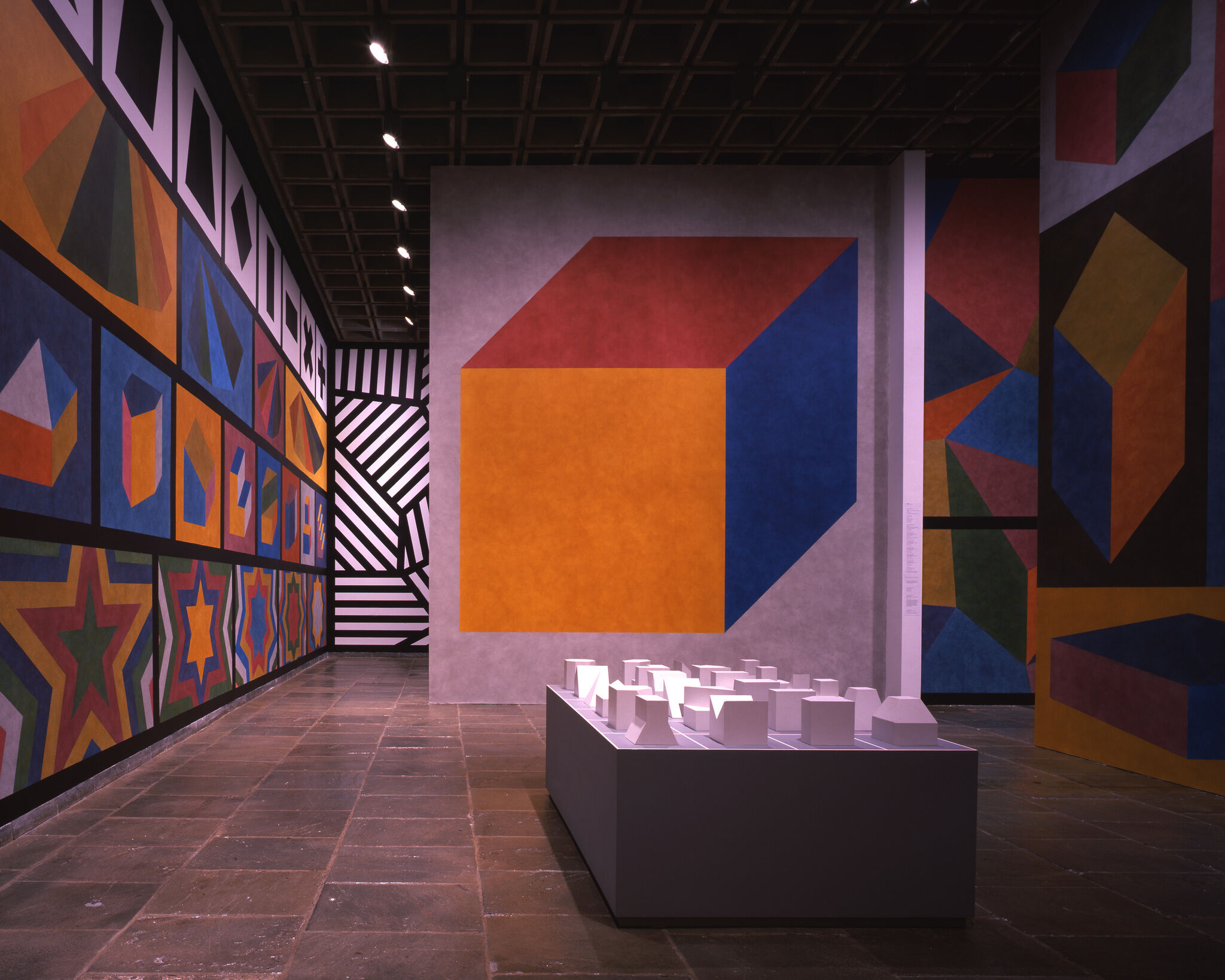 A gallery with walls covered in colorful geometric works of art and a display of small white geometric sculptures.