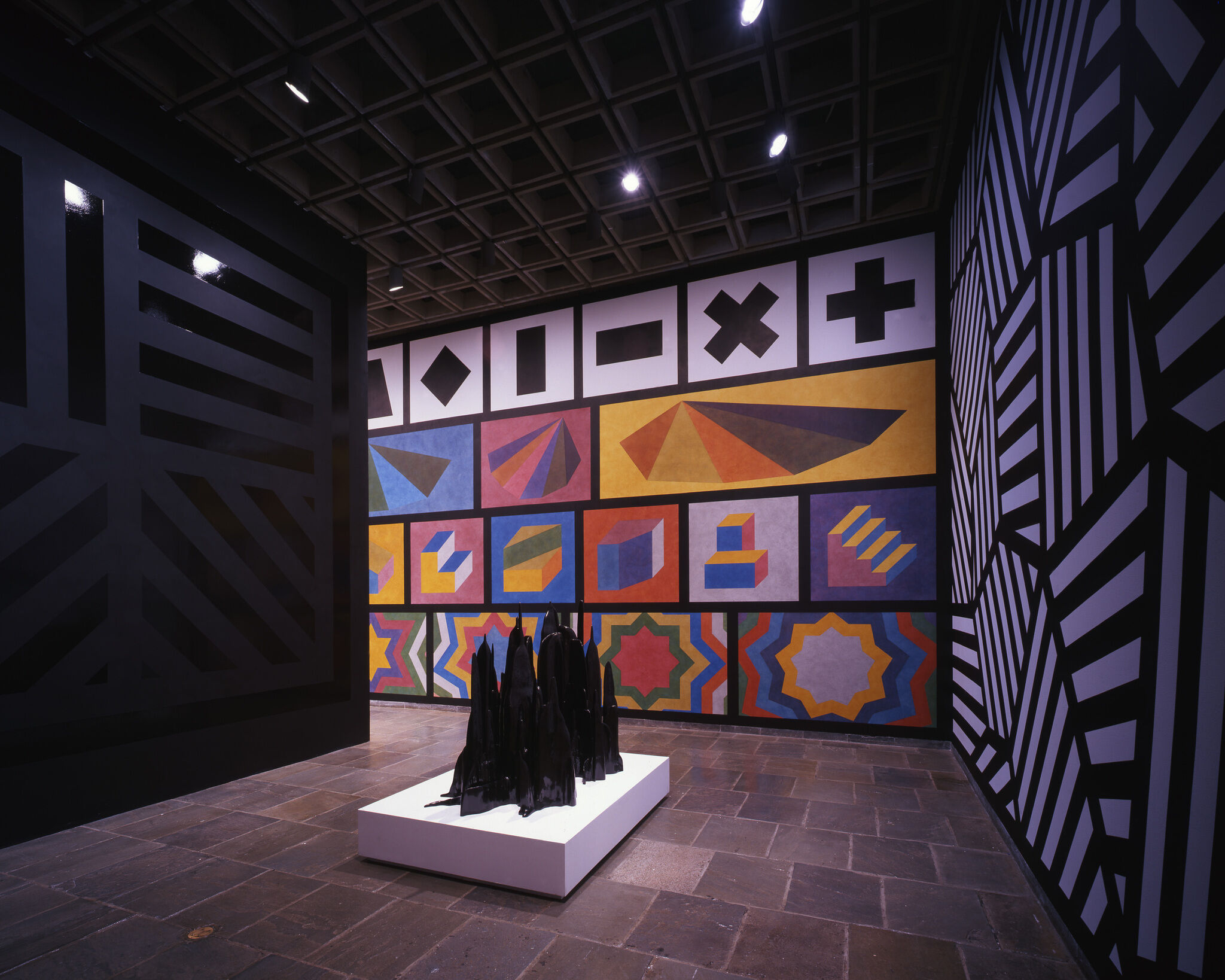 A gallery with two walls covered in black and white geometric patterns and another with colorful geometric figures, along with a black sculpture on display.