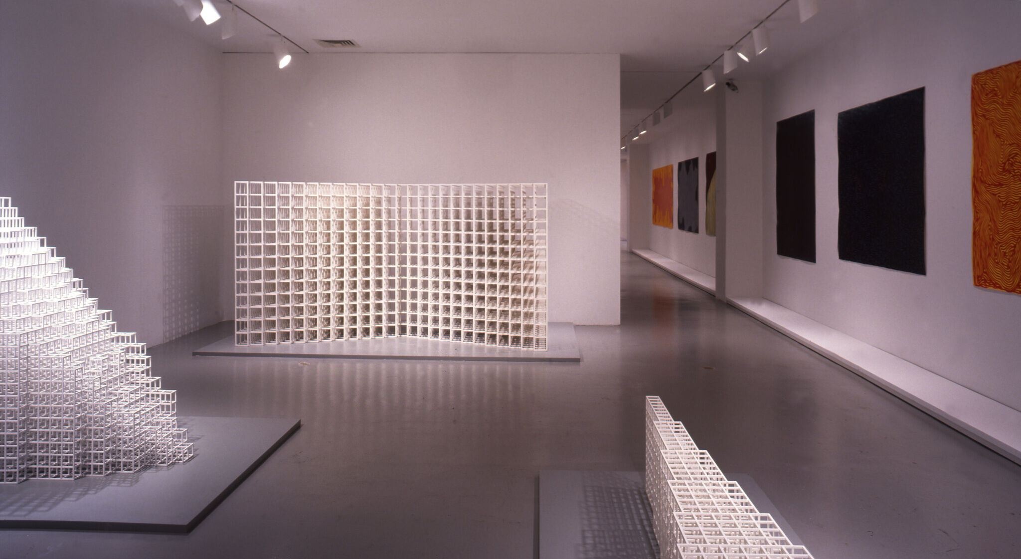 White sculptures made out of grids, along with abstract paintings displayed in a gallery.