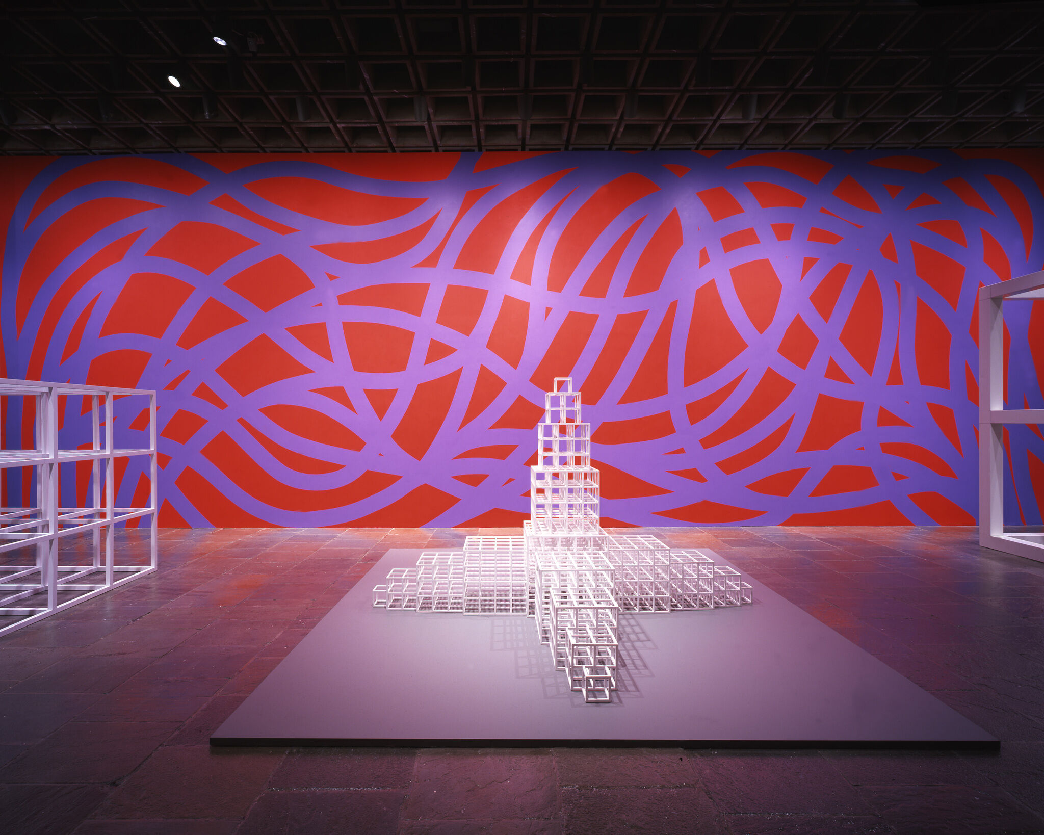 White geometric sculptures displayed in front of a red and blue abstract wall art in a gallery.