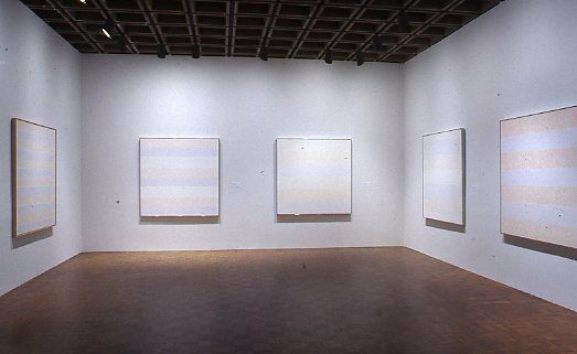 Square abstract paintings depicting faint stripes displayed in a gallery.