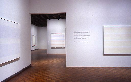 Abstract paintings depicting faint horizontal stripes, alongside wall text, displayed in a gallery.