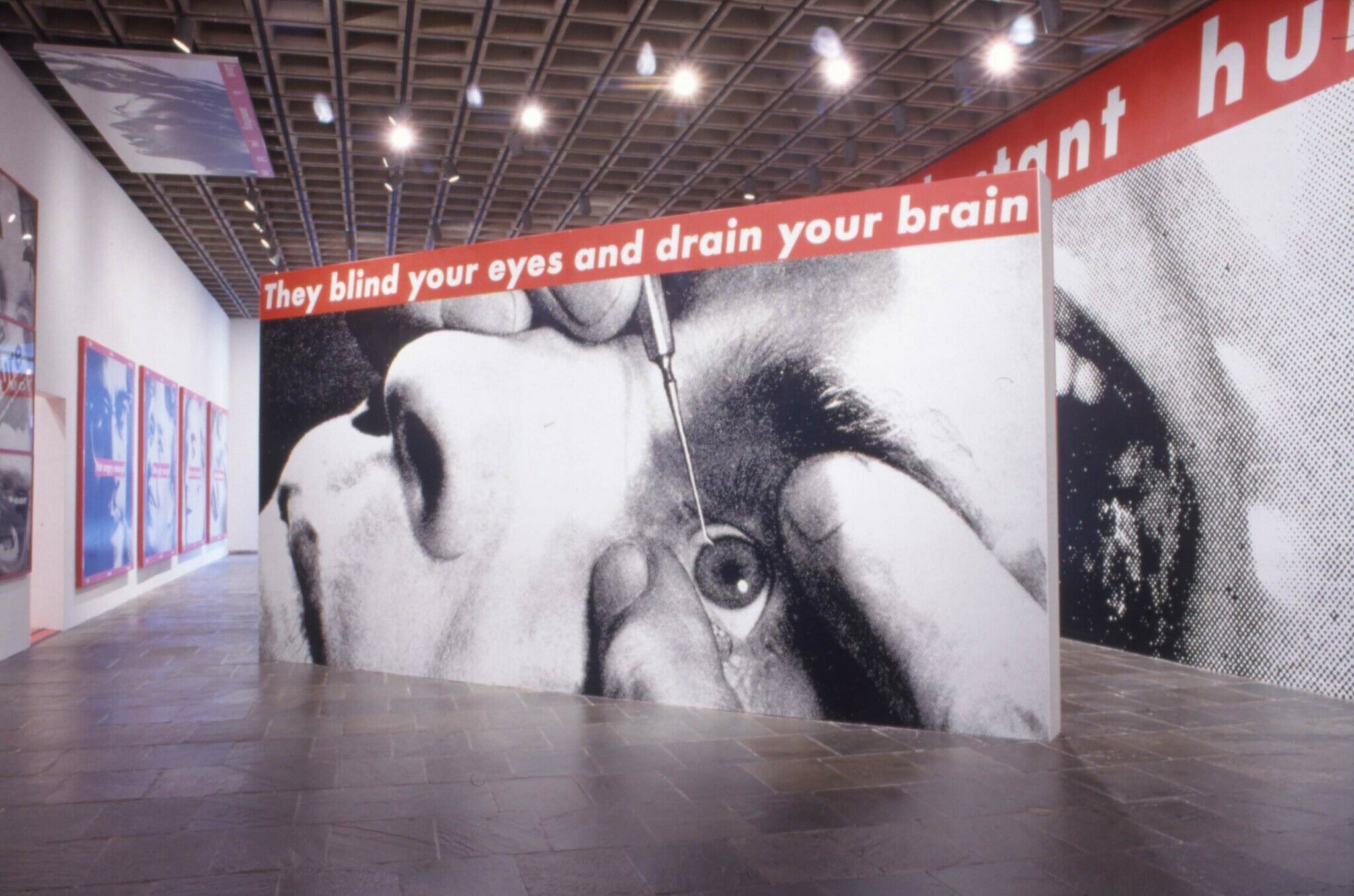 A wall in a gallery covered with a black and white photograph of a face with a tool pointed towards the eye.