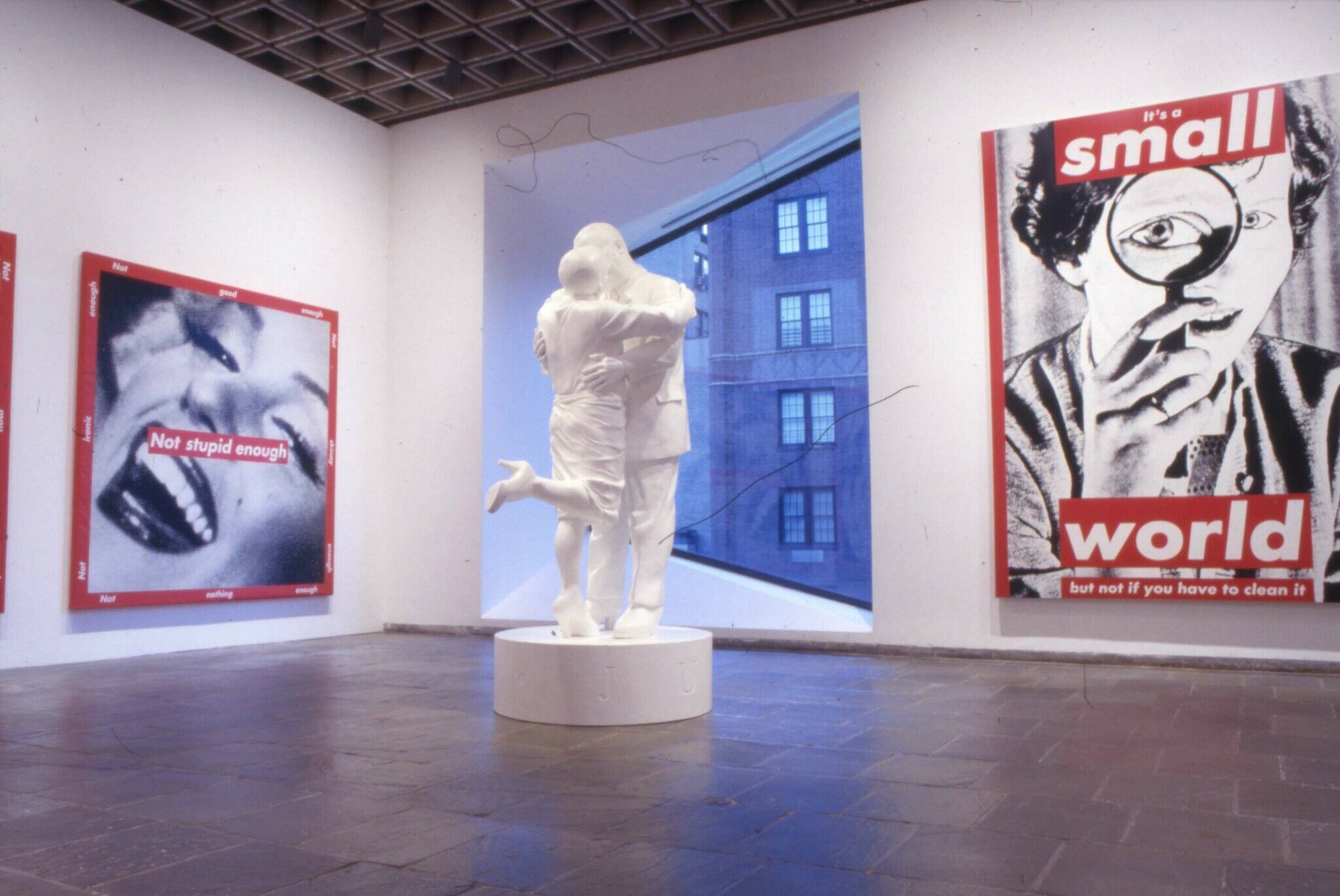 A white sculpture of two figures kissing, displayed in a gallery along with other works of art.