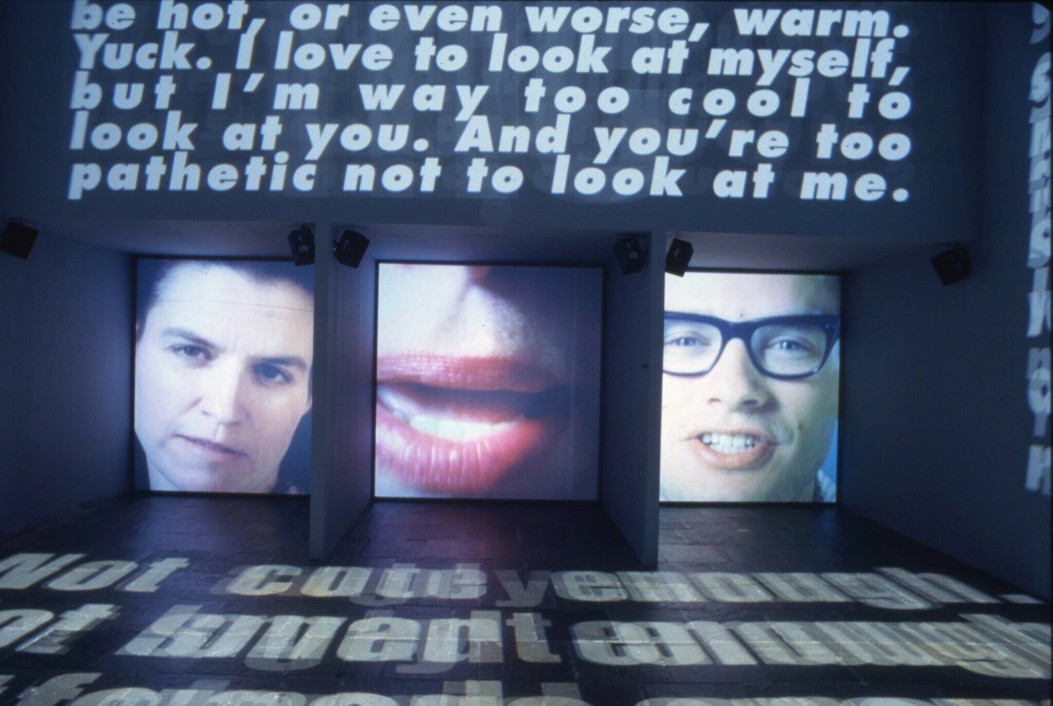 Three screens displaying close up views of a face, along with projections of white text on the walls.