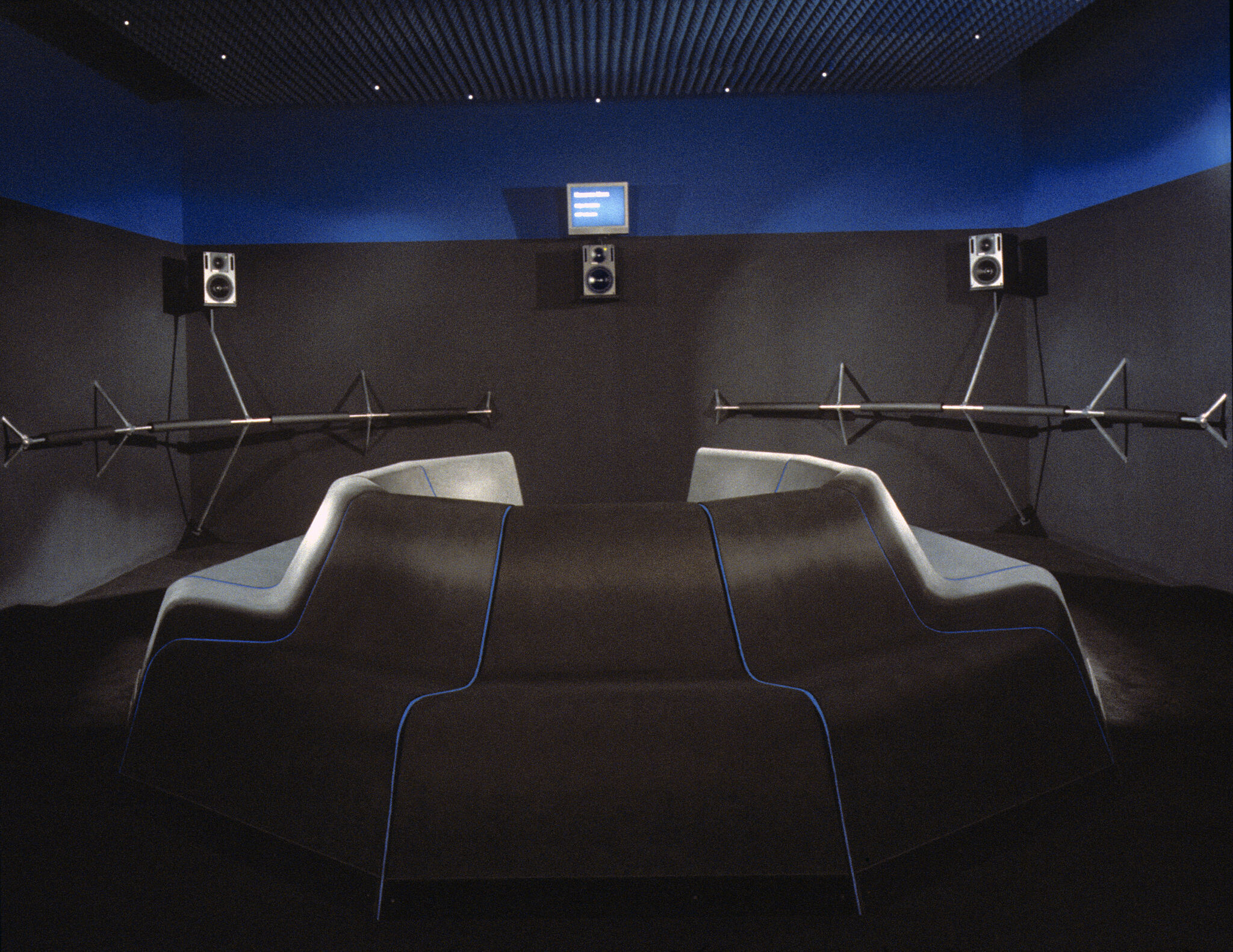 An installation view of a gallery with circular seating, dark gray walls, and three speakers.