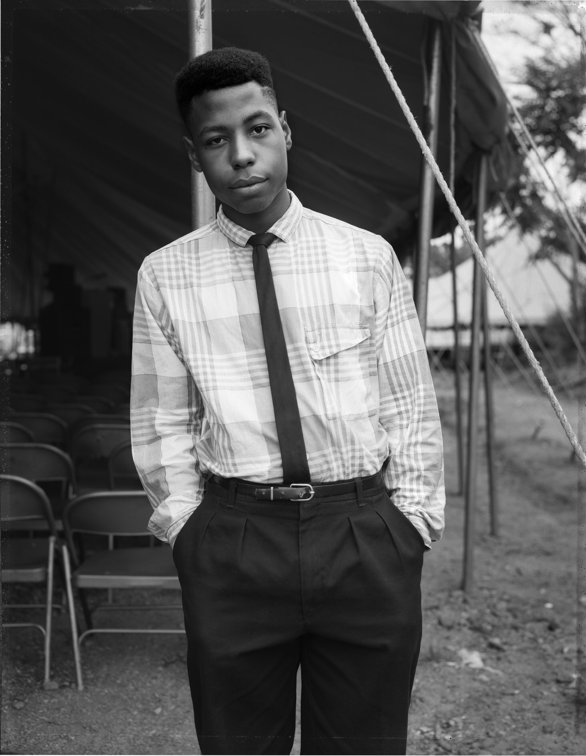 A young black man in formal attire stands in front of a tent and rows of metal chairs. 