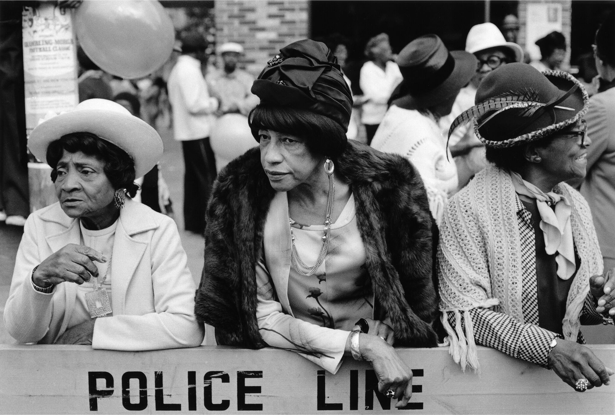 Three women in formal attire leaning against a parade barricade. 