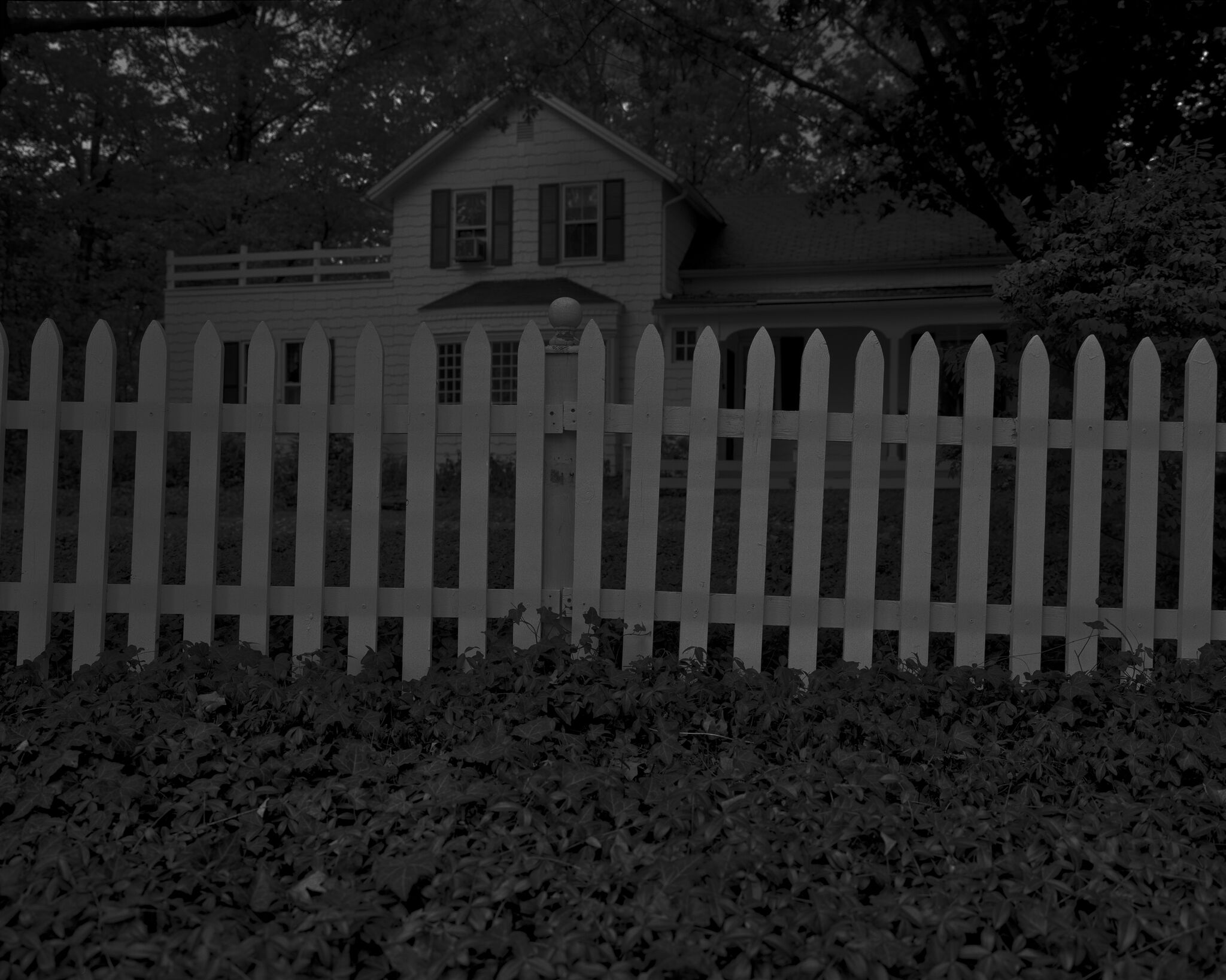 A greyscale close-up of a picket fence in front of a large house. 