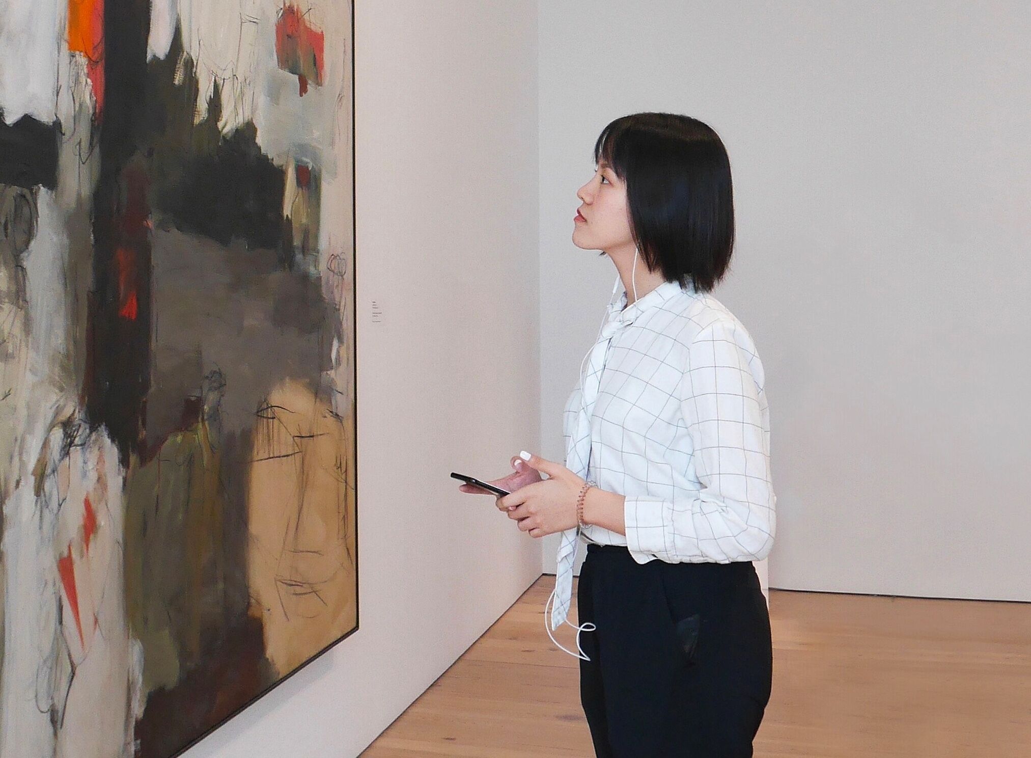 A woman viewing a work of art while wearing headphones and holding a smartphone.