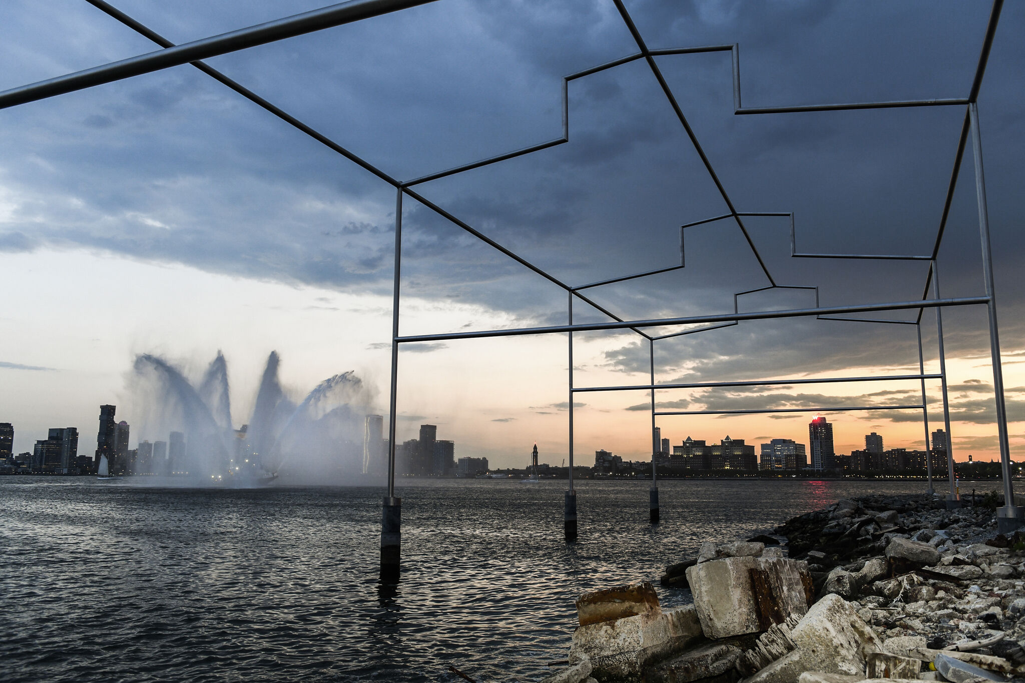 Close-up view of a sculpture shaped like the metal outline of a building on the shore of the Hudson River, with the New Jersey skyline visible in the background at sunset, and a fountain-spraying boat in the distance.