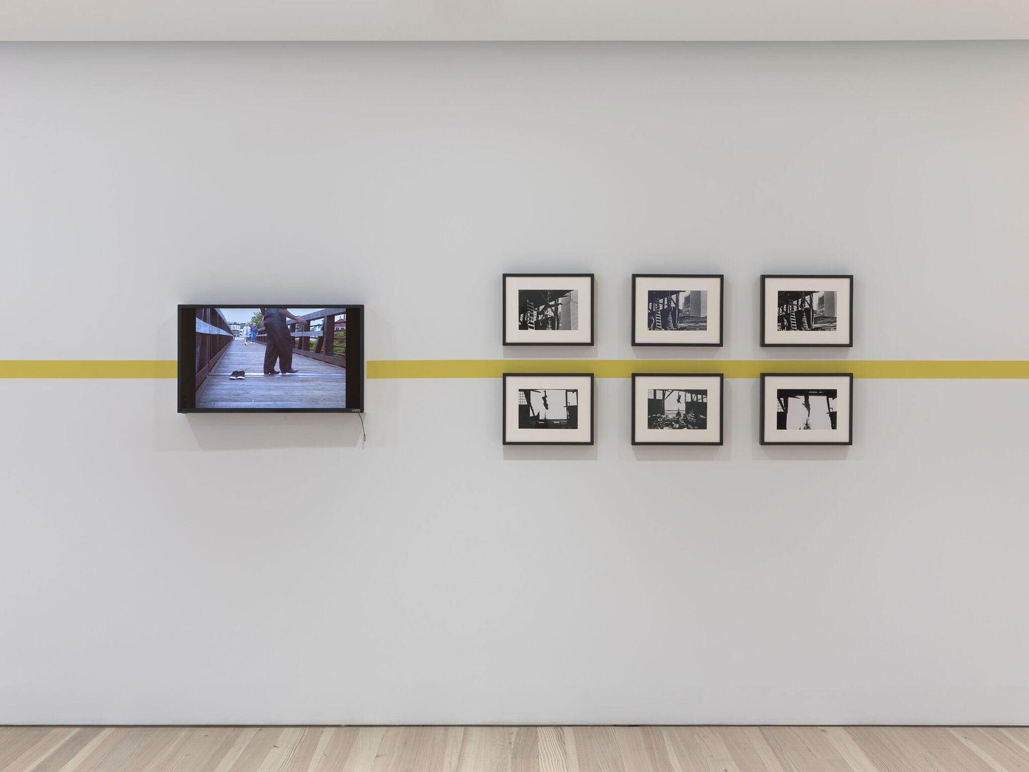 A black screen and a series of six framed photographs are mounted against a white exhibition wall.