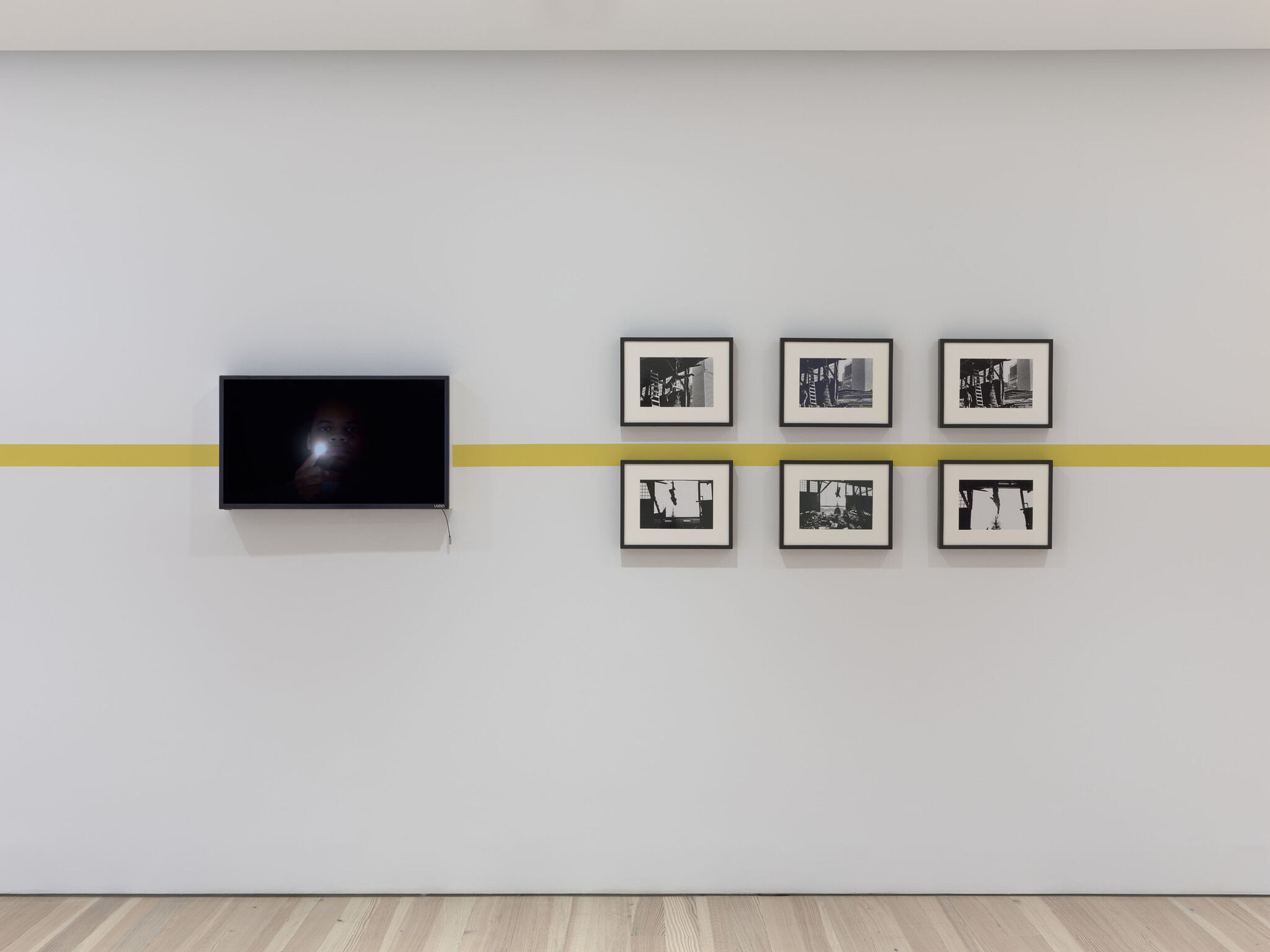 A black screen and a series of six framed photographs are mounted against a white exhibition wall.