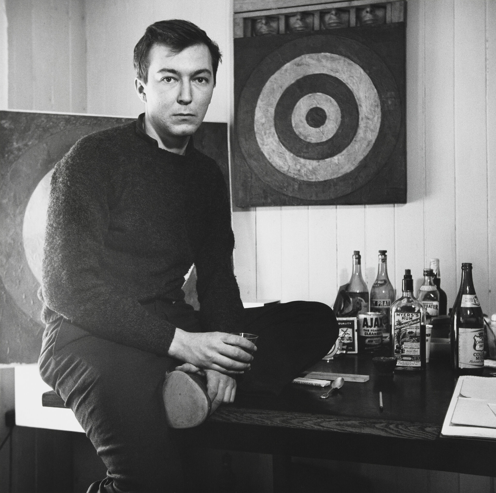 A young man sitting on the edge of a desk, looking at the viewer with a neutral expression, and holding a glass, against a backdrop of a painting on the wall and a collection of liquor bottles.