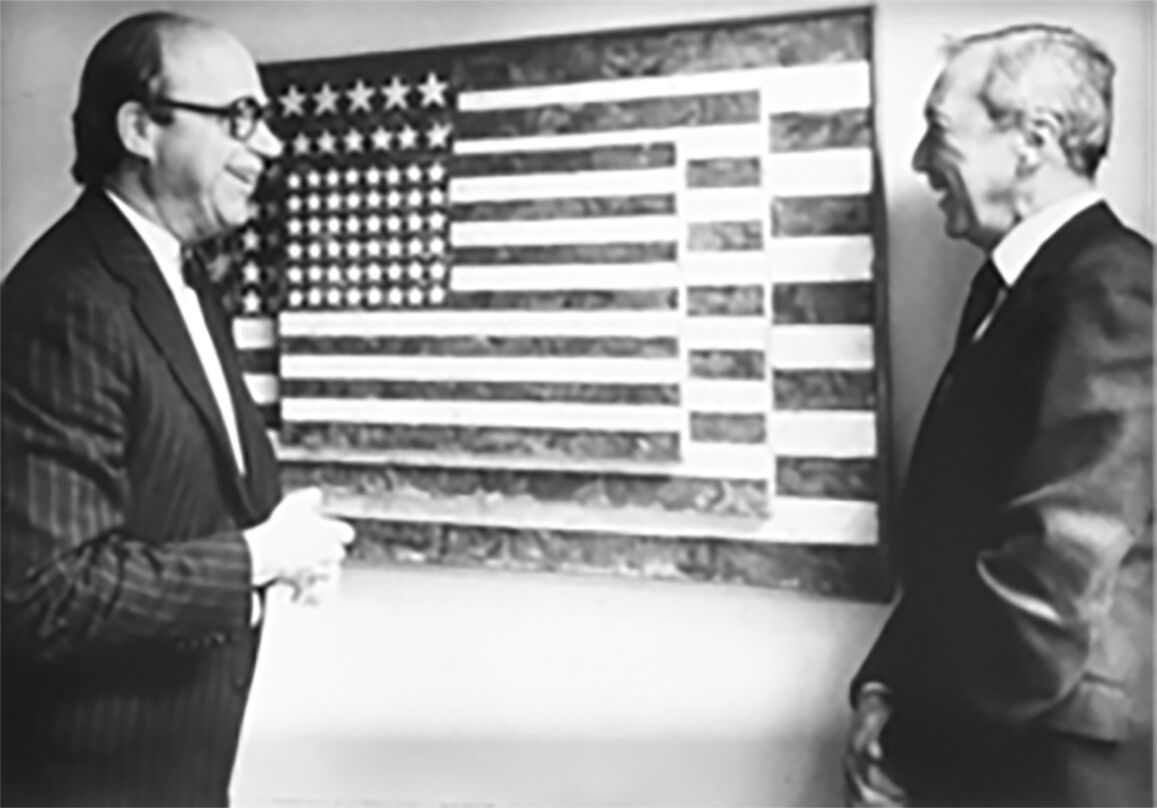 Two men standing facing each other, with a painting of an American flag on the wall behind them.