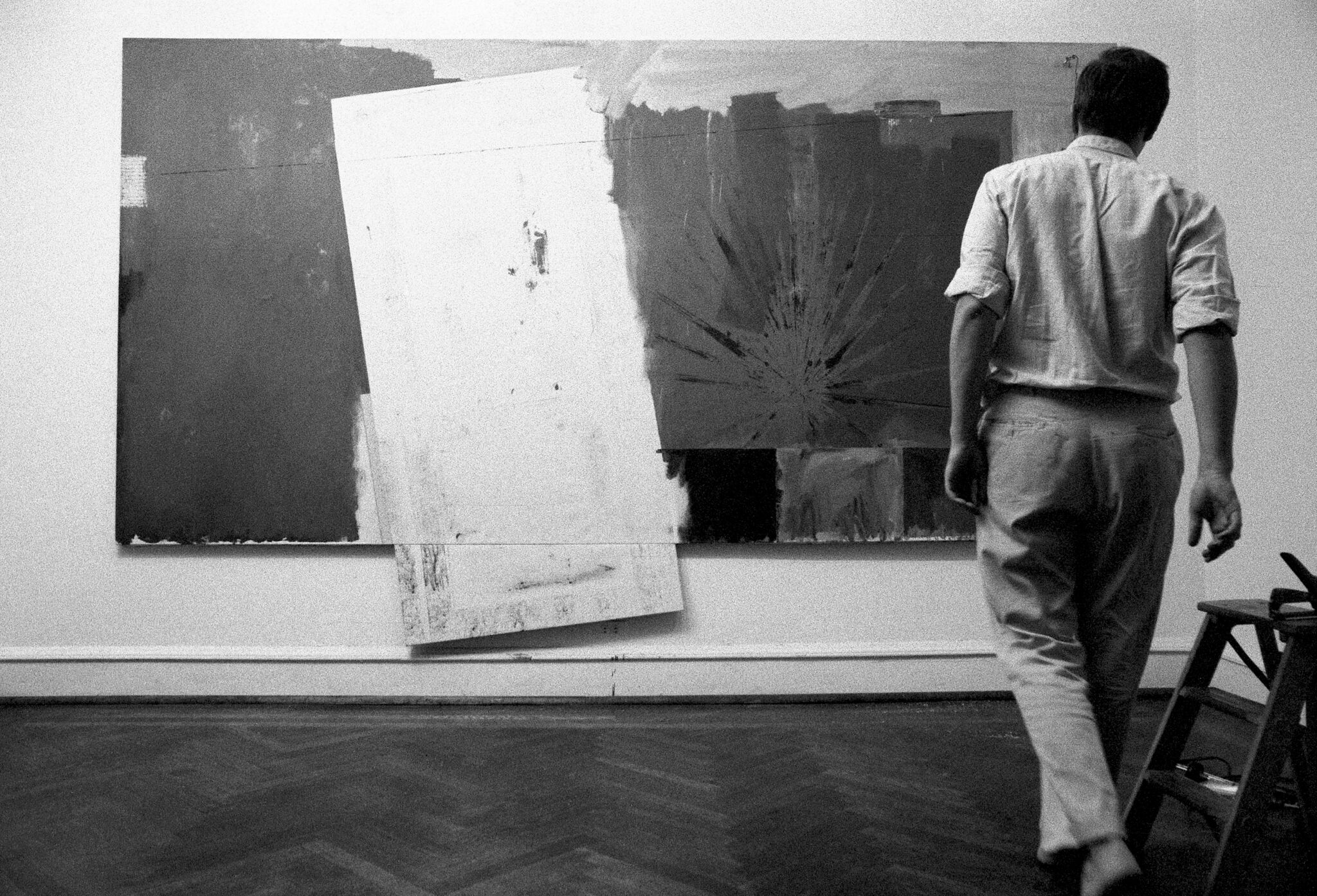 Jasper Johns stands facing away from the camera looking at one of his works mounted on the wall. 