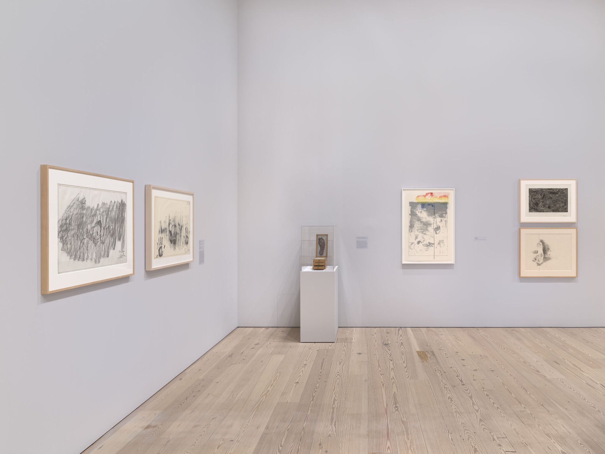 A white exhibition room with five framed works and a sixth work encased in glass.
