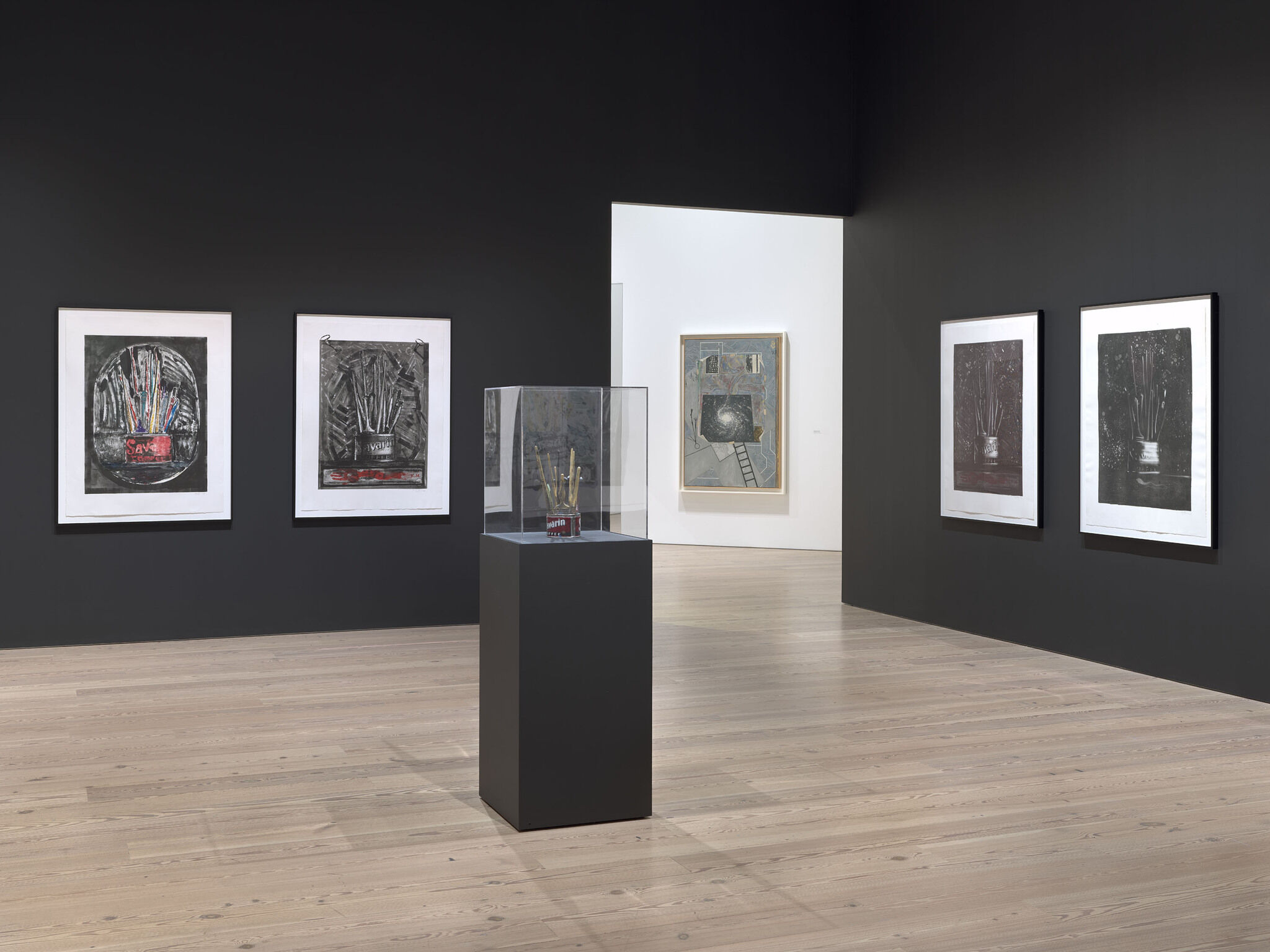 A black and white exhibition room with five framed works mounted on the walls and a sixth encased work in the center of the room.