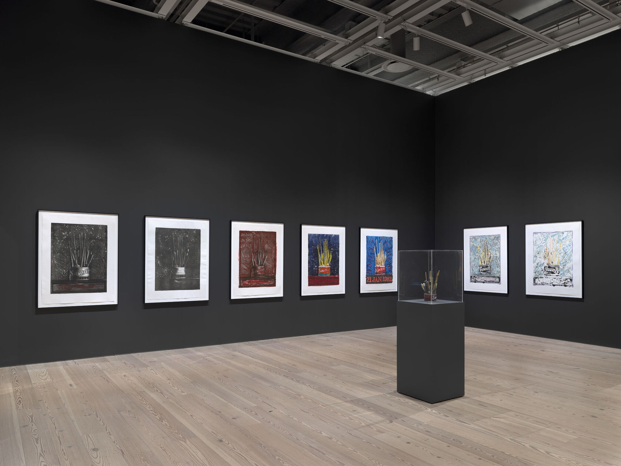 A black exhibition room with seven framed works mounted on the walls and an eighth work encased in glass in the center of the room.