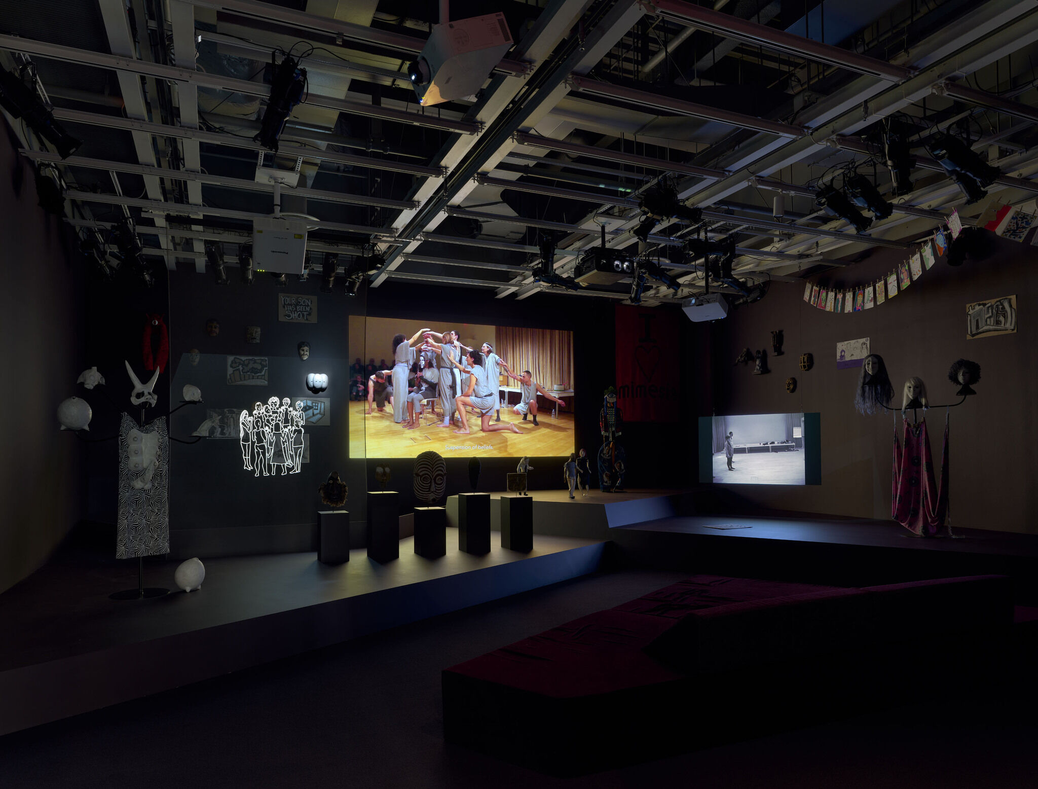 A dark exhibition room featuring a large digital screen and a series of smaller pieces installed throughout.