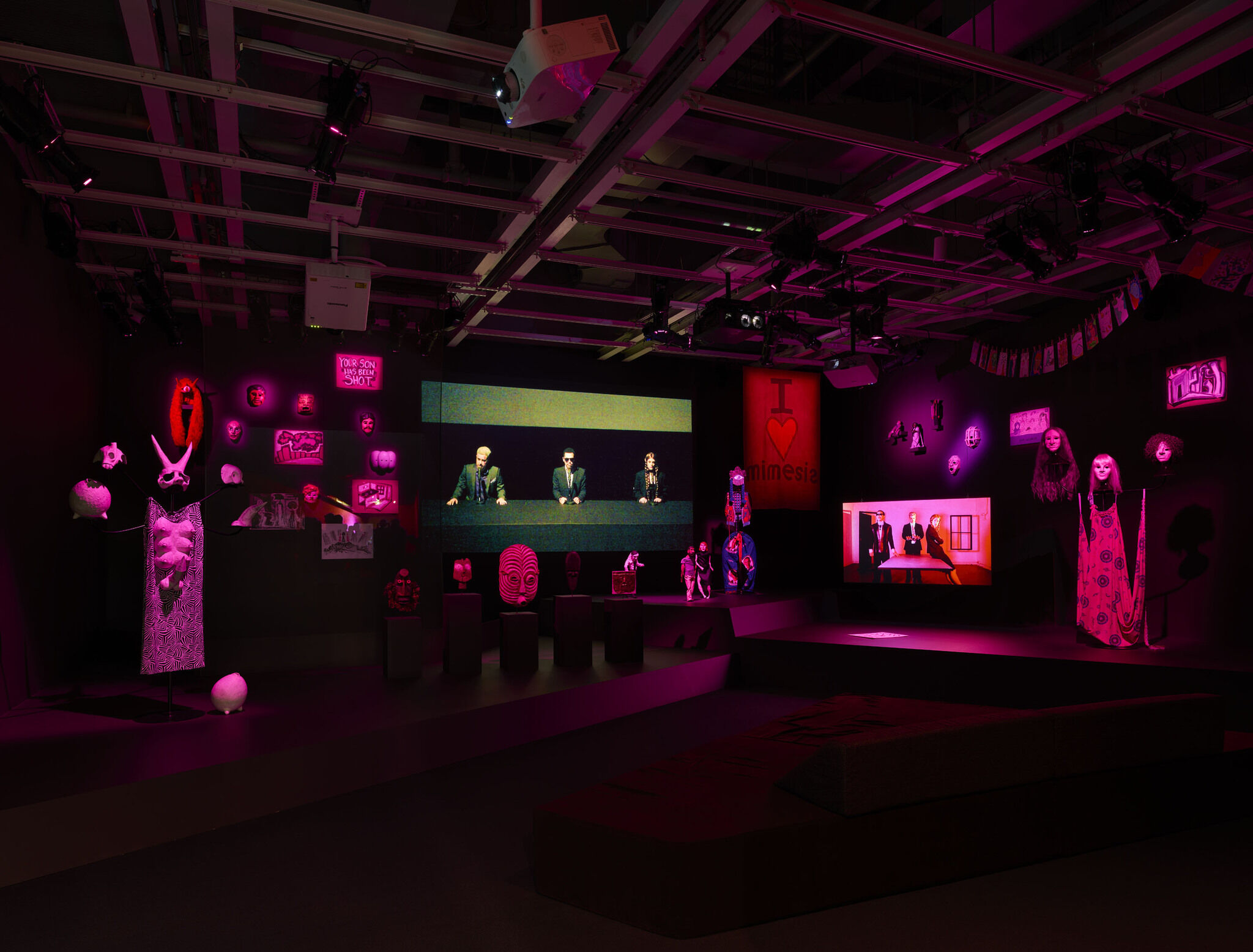 A dark exhibition room featuring a large digital screen and a series of smaller pieces installed throughout.