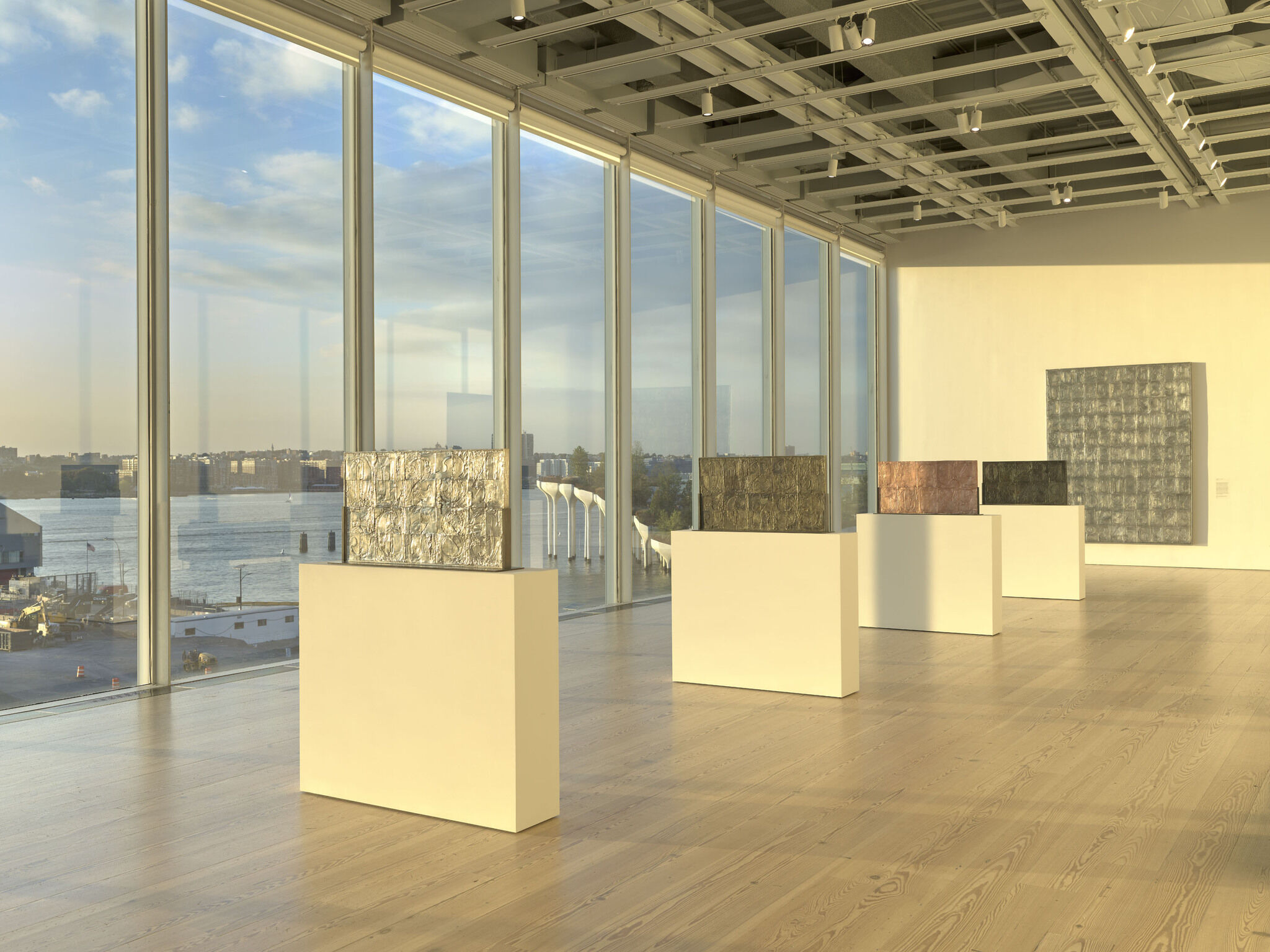 A white exhibition room with a wall of glass windows and a series of works installed down the middle of the room.