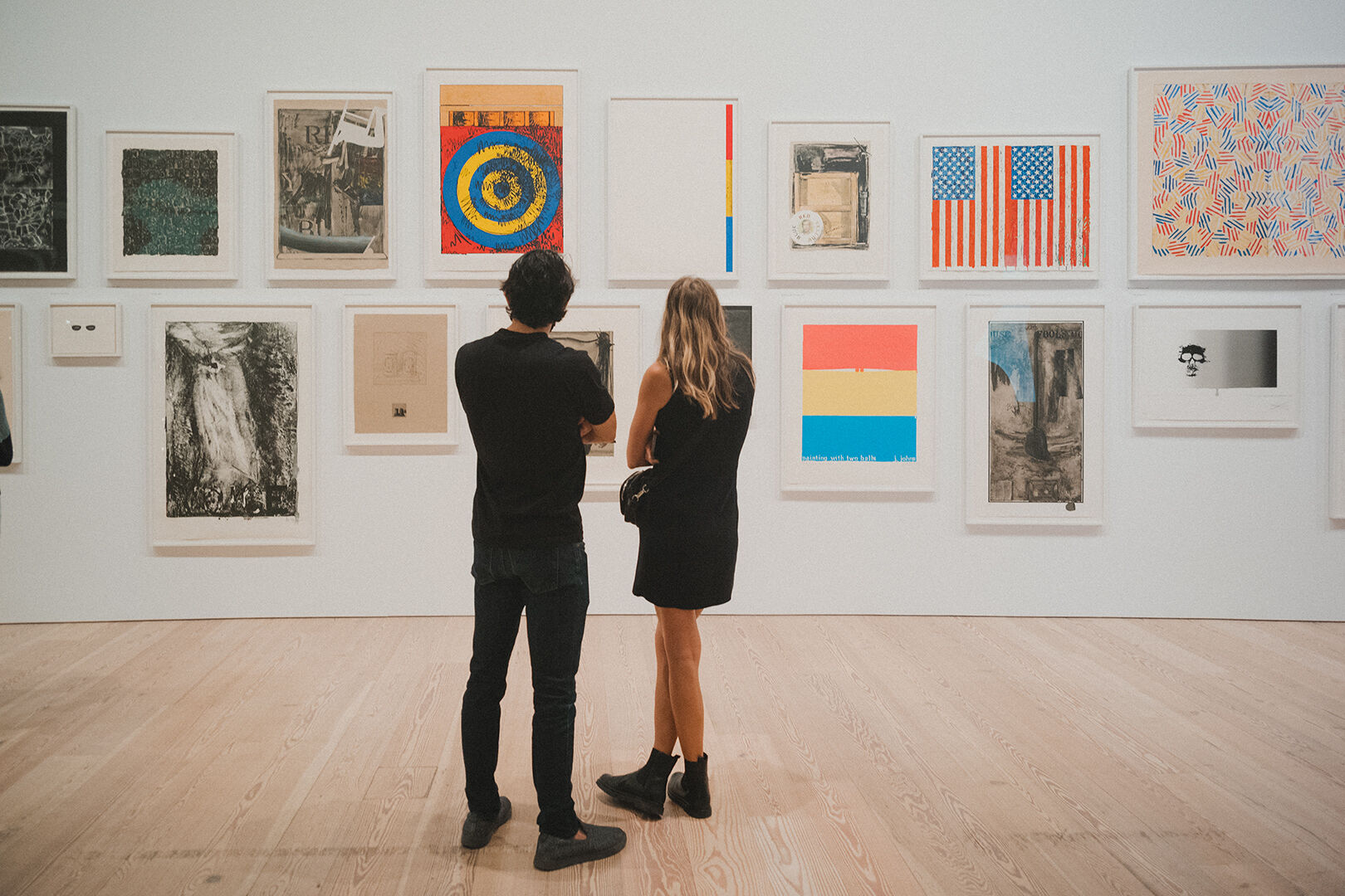 Two people standing in front of a white exhibition wall featuring a series of framed works. 