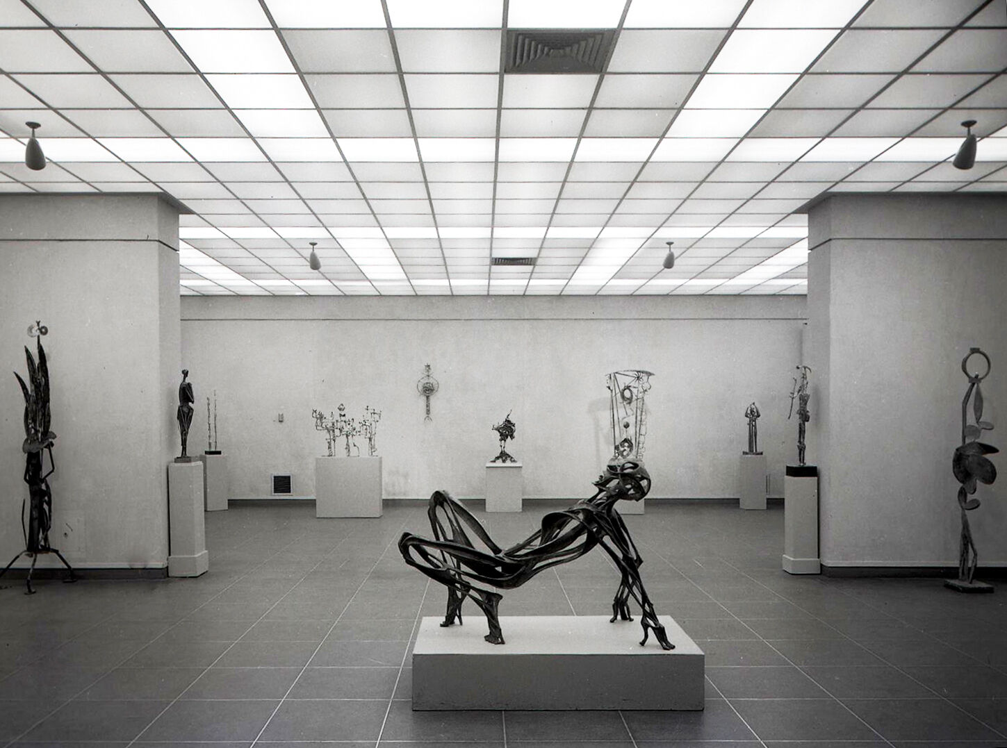 Black-and-white photograph of gallery of sculptures, with a central sculpture of a human figure in the foreground.