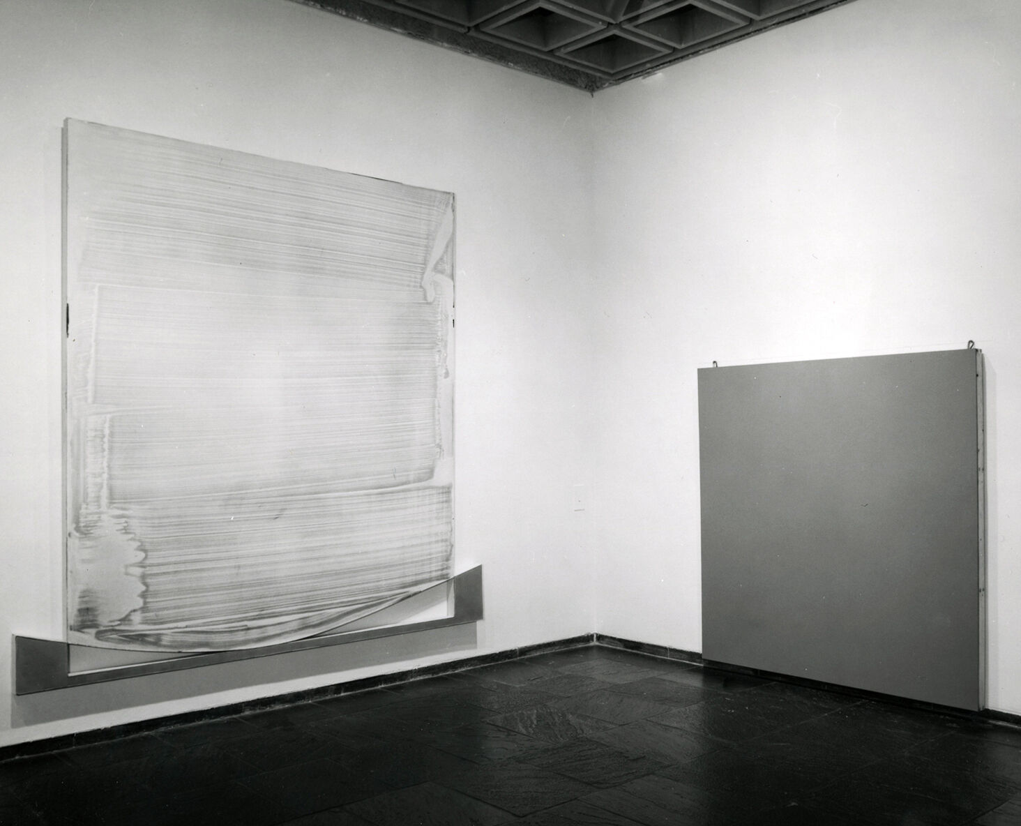 Black-and-white photograph of two paintings hanging in the corner of a gallery.