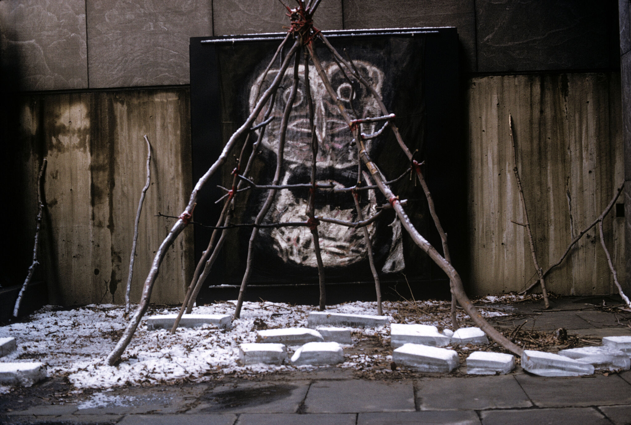 Tent of sticks and paving stones in front of an image of an owl-like figure.