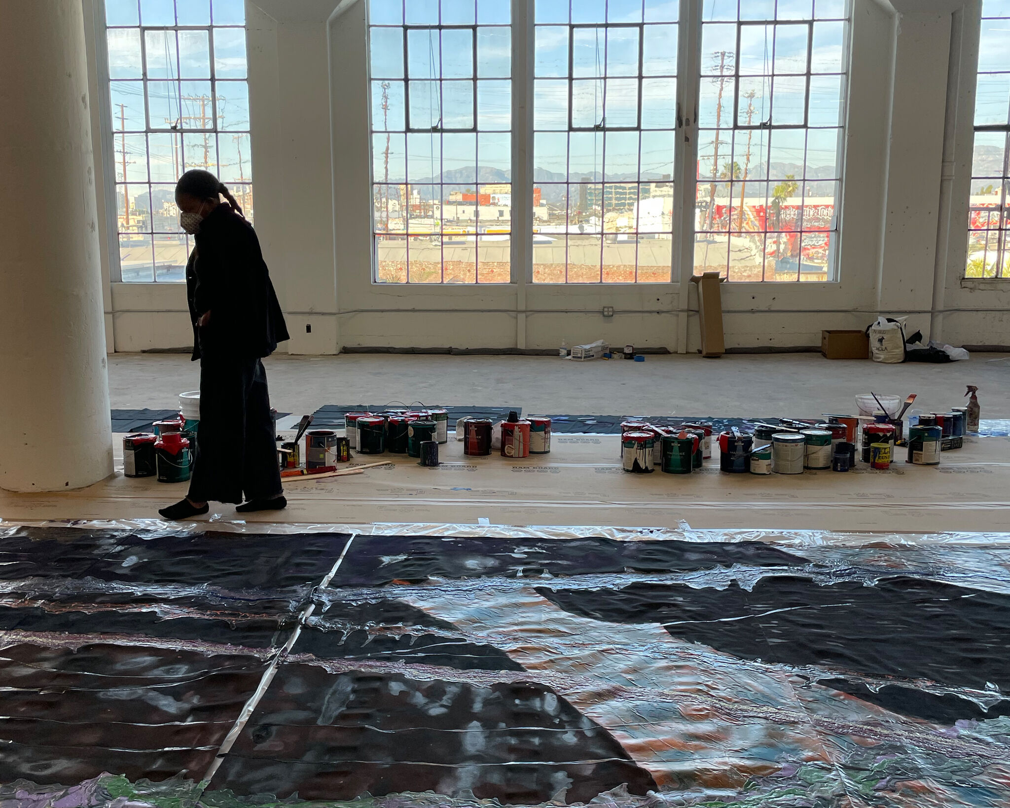 A woman stands surveying a large work of art that is laid out on the floor, in front of a bay of windows.