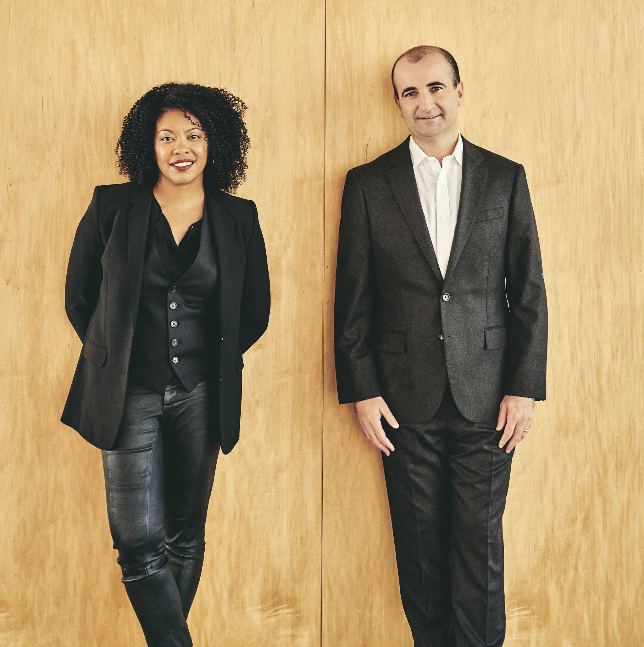 Curators Adrienne Edwards and David Breslin in all-black ensembles leaning against a wooden wall