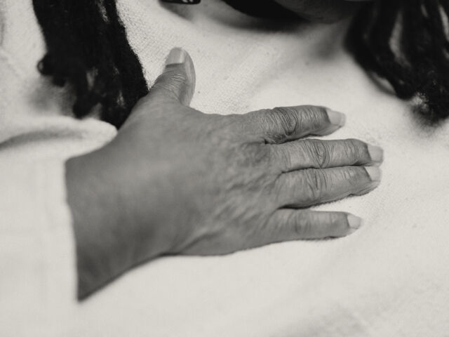 A black and white image of a Black woman's hand resting on her chest. She wears white long-sleeved shirt.