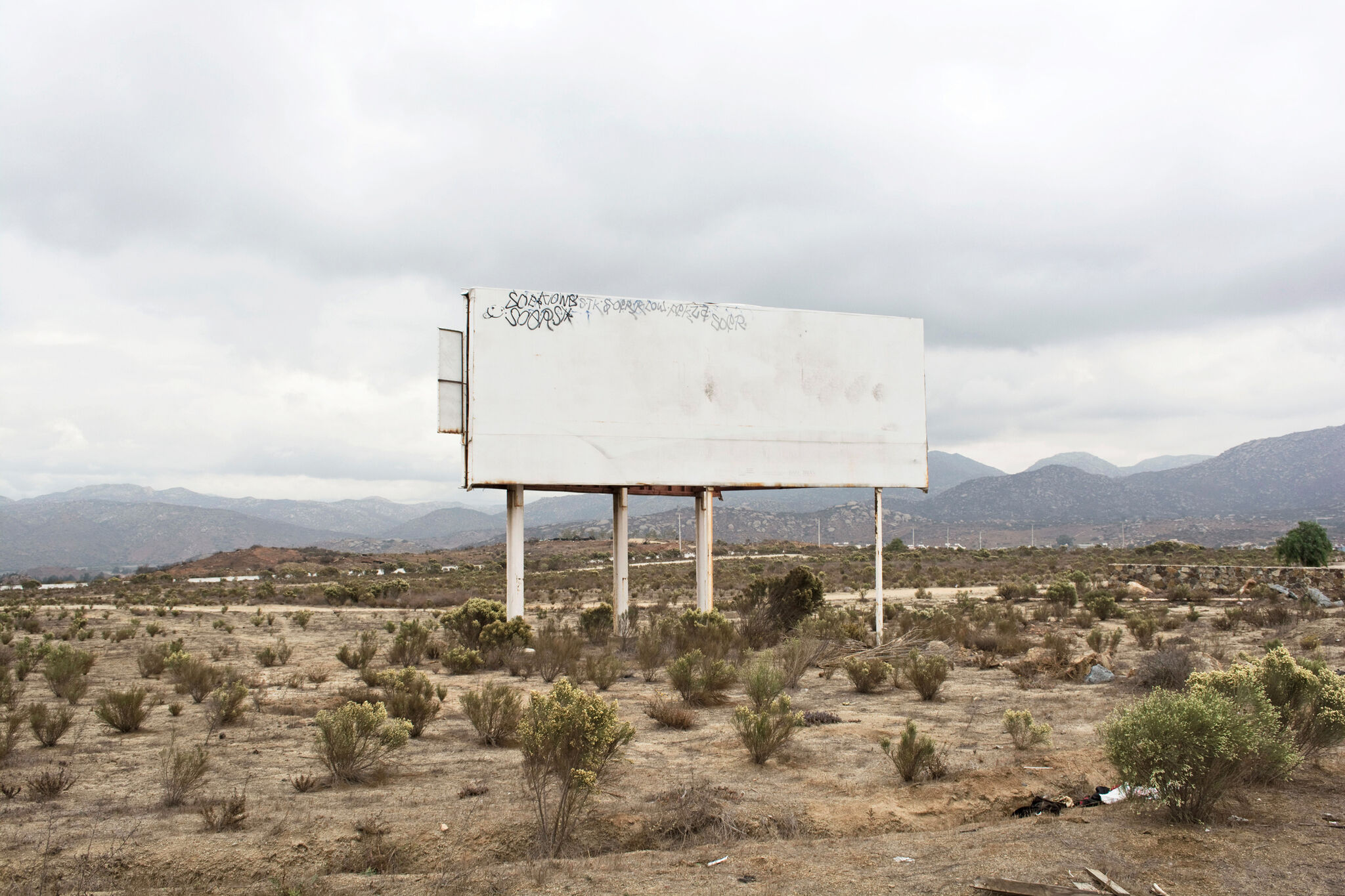 Photograph of a blank, white billboard with some graffiti in its upper left corner, set in a desert landscape with a gray sky and mountains in the background.