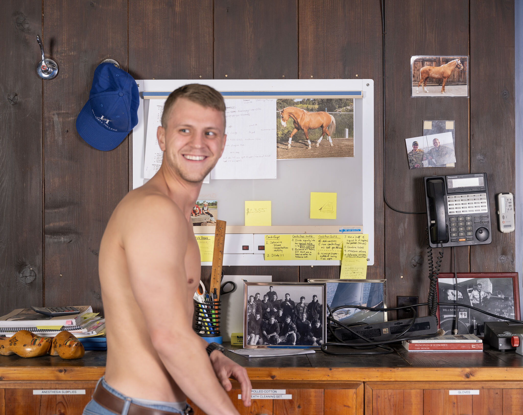 A smiling, shirtless, blonde man glances sideways out of the frame while standing in front of a wood-paneled office space that is cluttered with a bulletin board, pinned and framed photographs, a wall-mounted phone, sticky notes, and various office supplies.