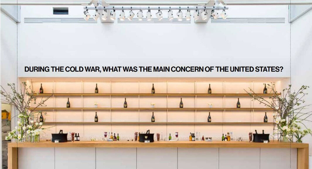 A line of screenprinted text in block capitals affixed to a wall above shelves and a bar. The text reads: "During the Cold War, what was the main concern of the United States?"