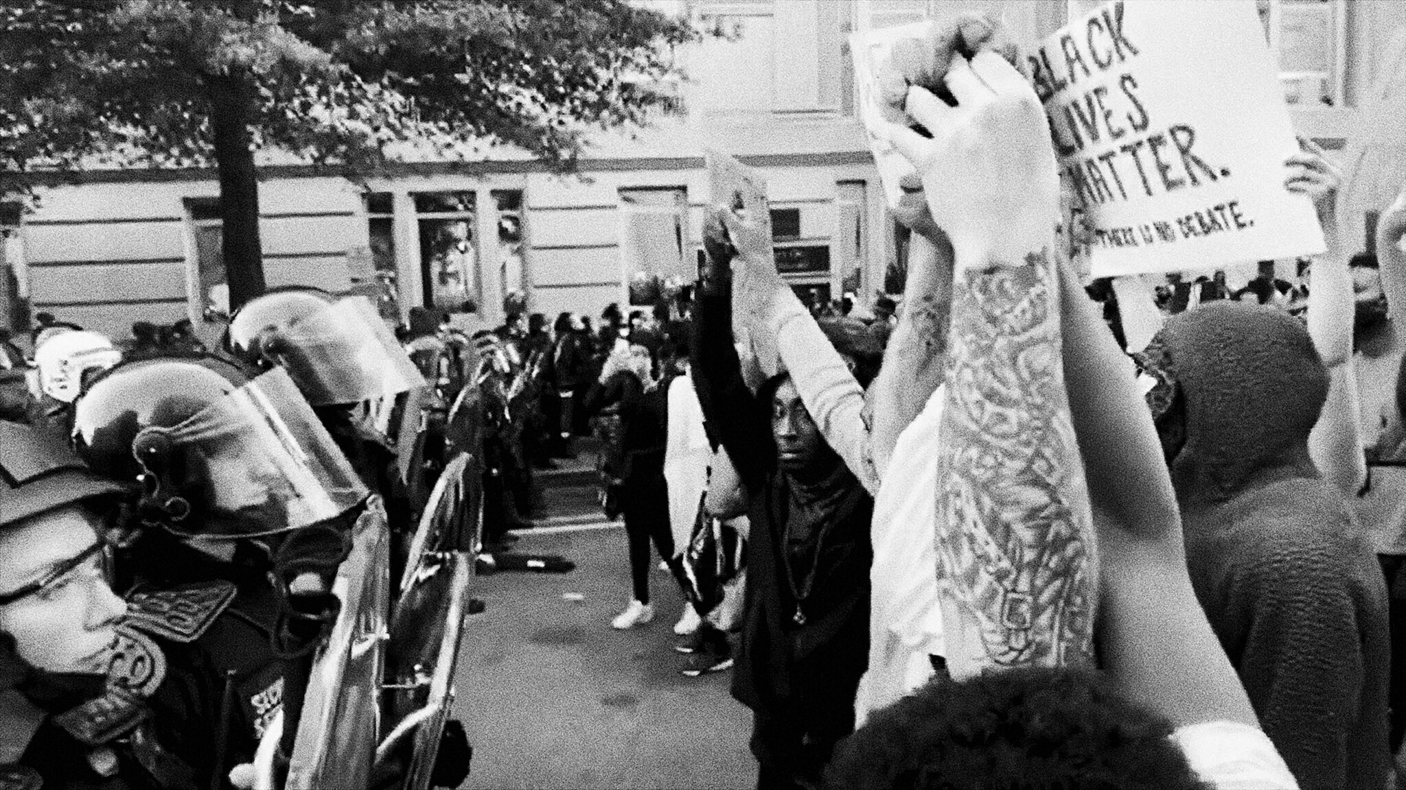 Black-and-white image of protesters holding up signs (one of which reads "Black Lives Matter. There is no debate.") facing off against a line of police officers wearing riot gear.