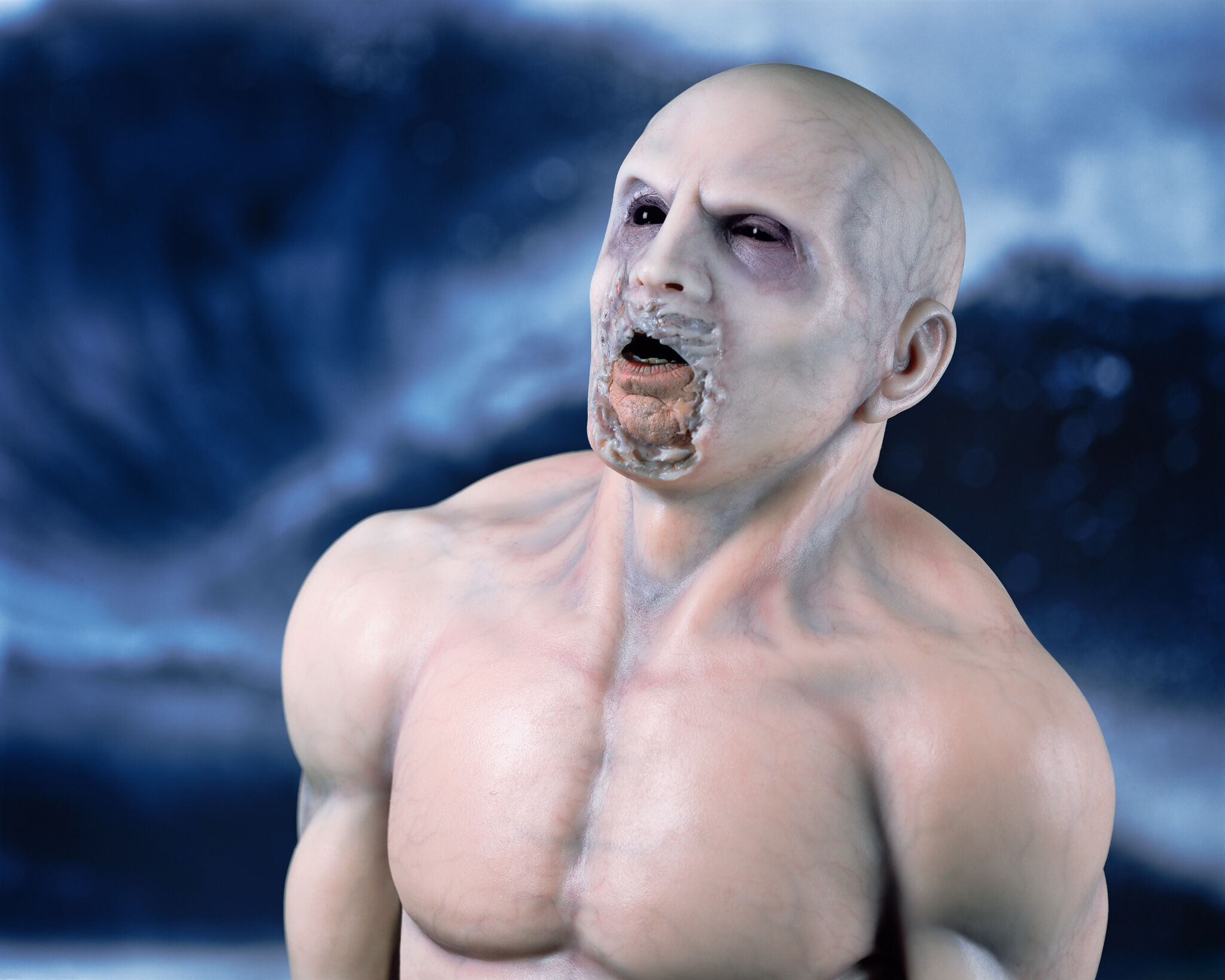A shirtless, bald, muscular, humanoid figure with black eyes and a disfigured, marled mouth, with blue veins showing through the skin.