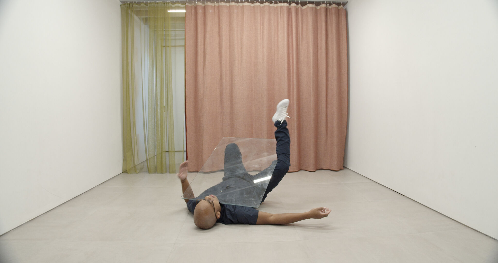 A person in a room is lying face-up on the floor with limbs akimbo and beneath a pane of glass.