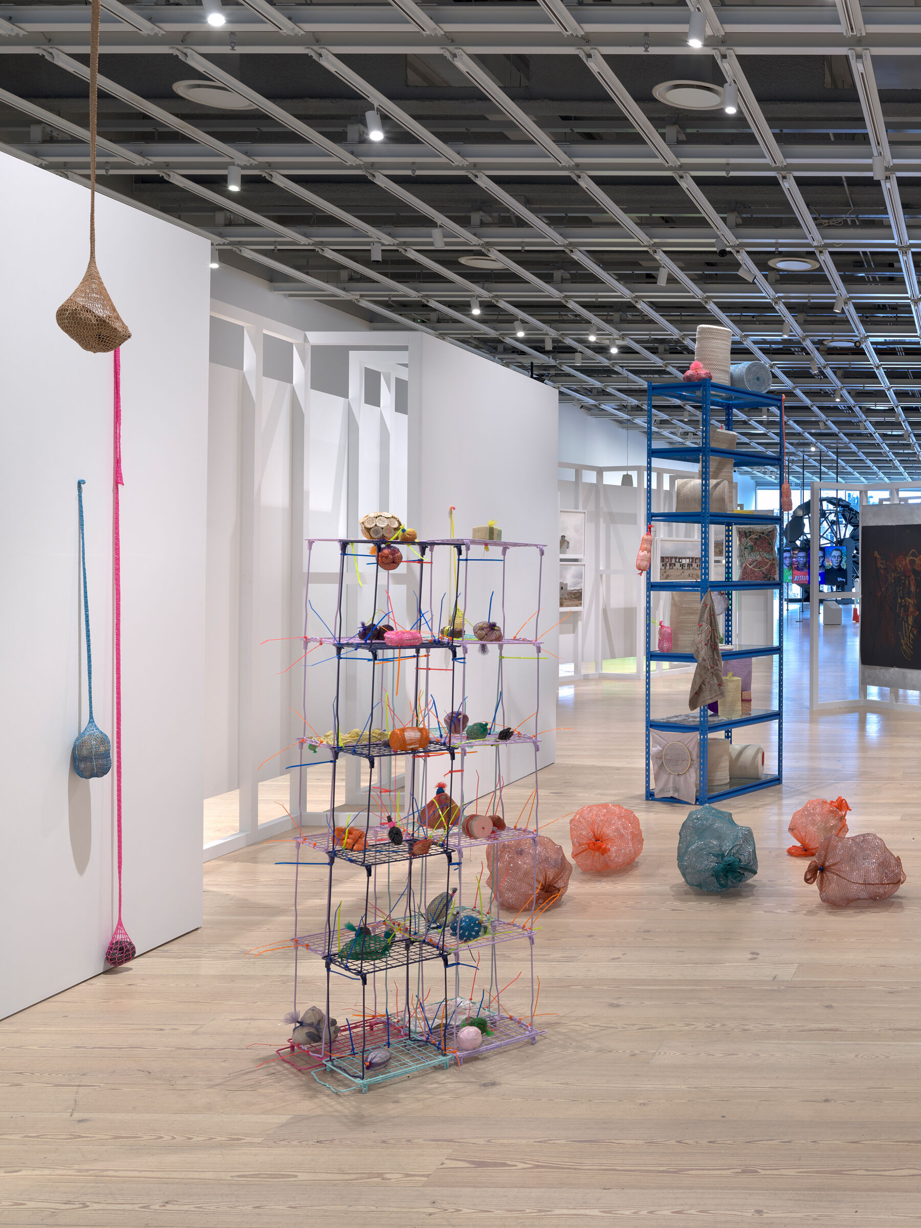 An installation view of an artwork consisting of colorful, indiscernible materials and objects presented on a wire shelving unit and hanging from the wall. 