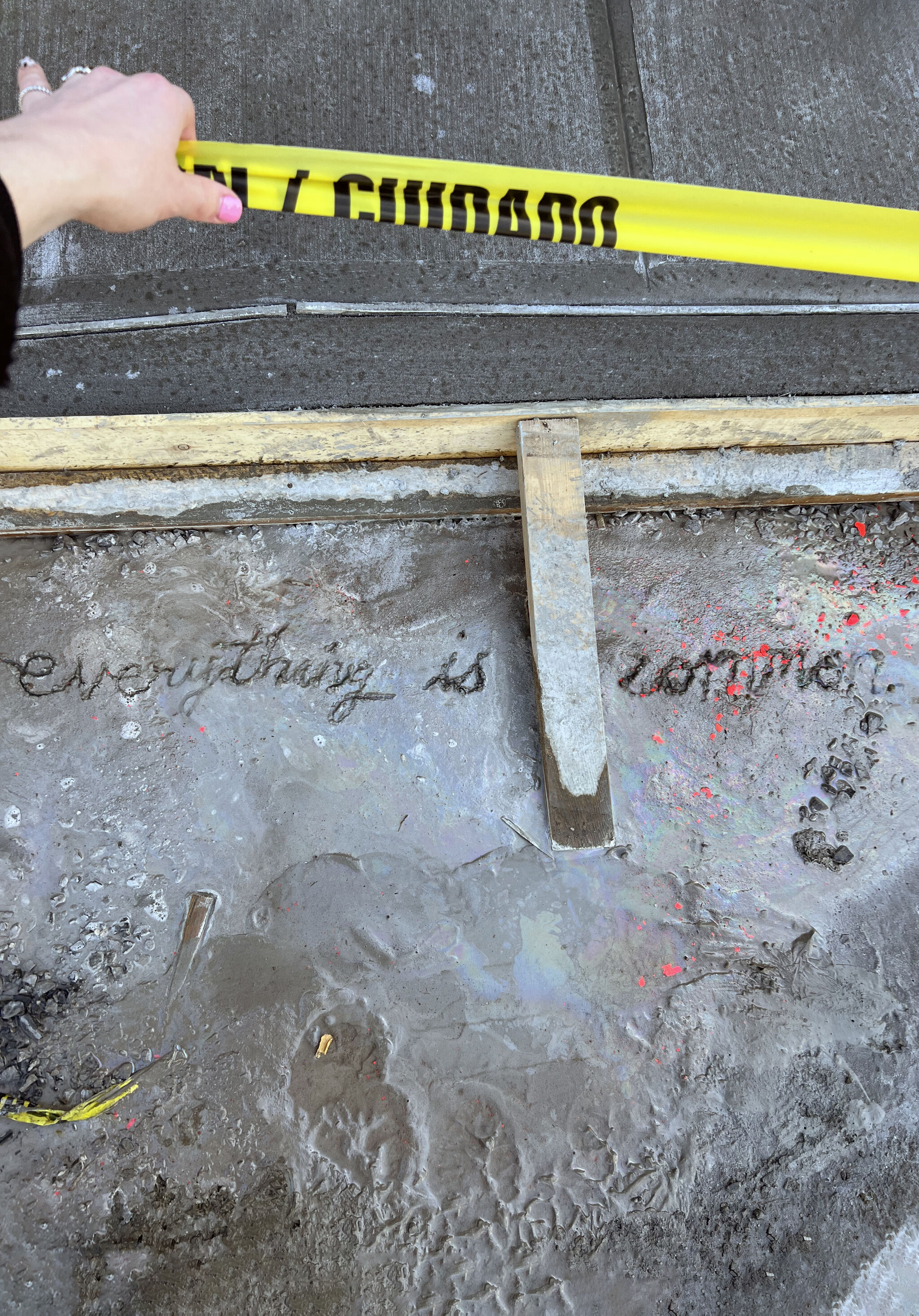A person holding caution tape over a patch of wet cement with the words Everything is Common written into it.