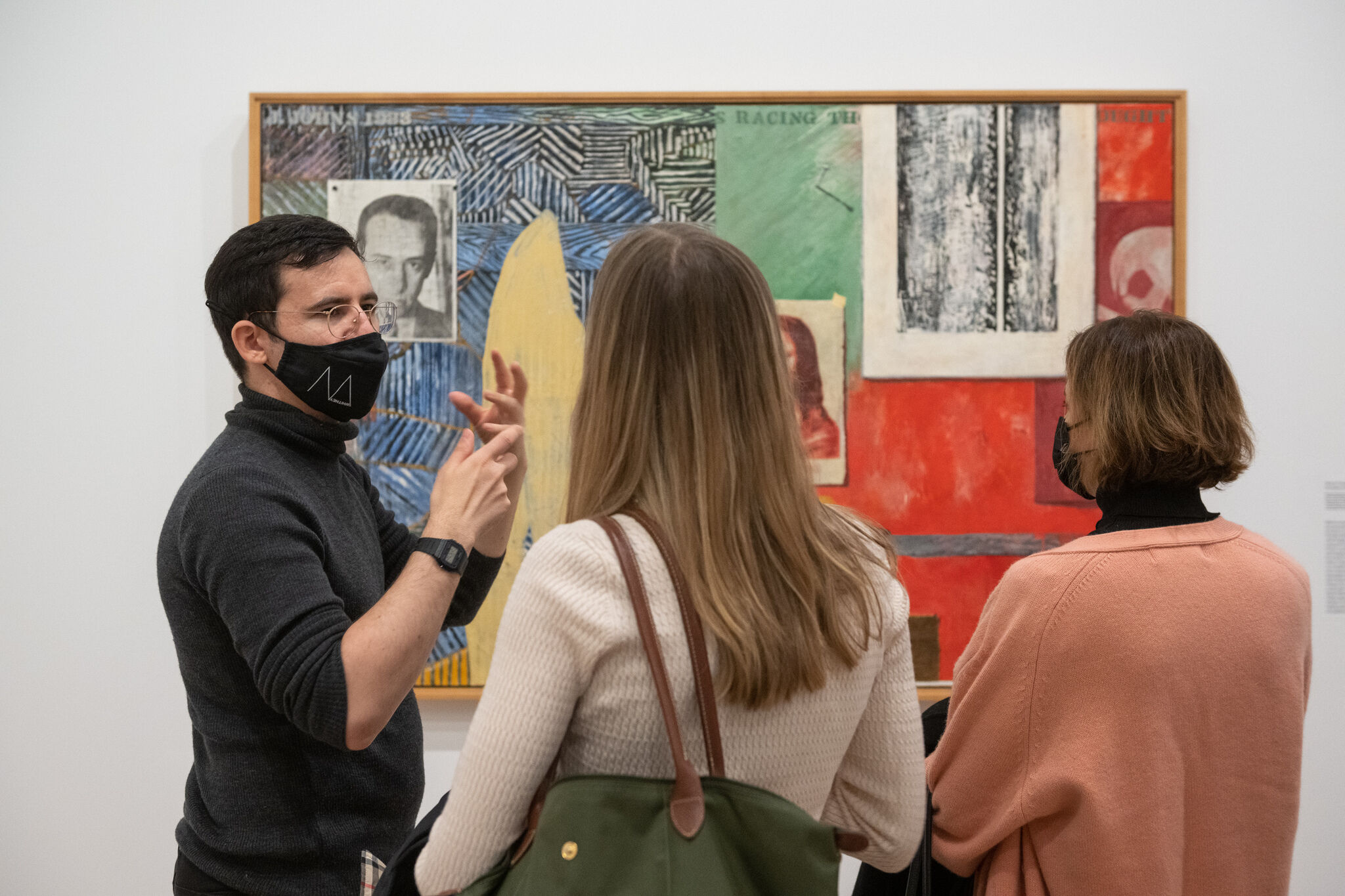 A Joan Tisch Teaching Fellow, wearing a black mask, talking to two visitors as they look at an installed artwork.