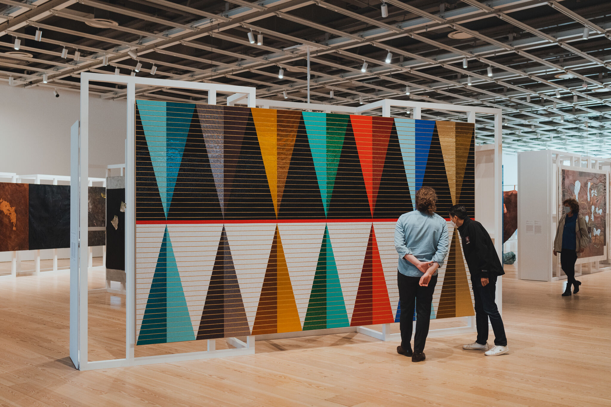 Three visitors walk through an open gallery space and inspect works of art, including a large canvas of a colorful triangle pattern.